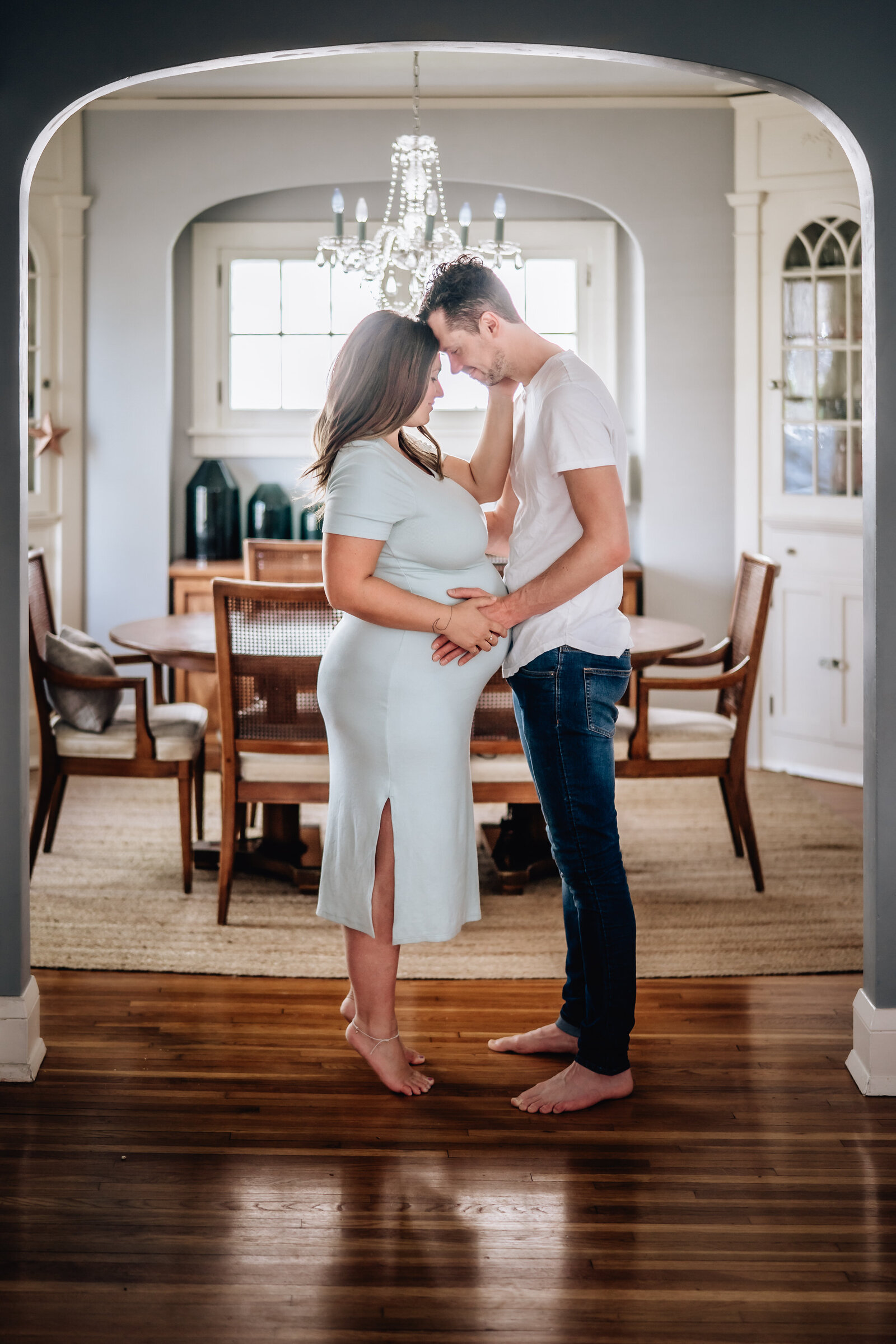 A pregnant couple stands barefoot in a warmly lit dining room, holding each other closely and touching foreheads. The woman is wearing a light blue dress, and the man is dressed casually in a white t-shirt and jeans.