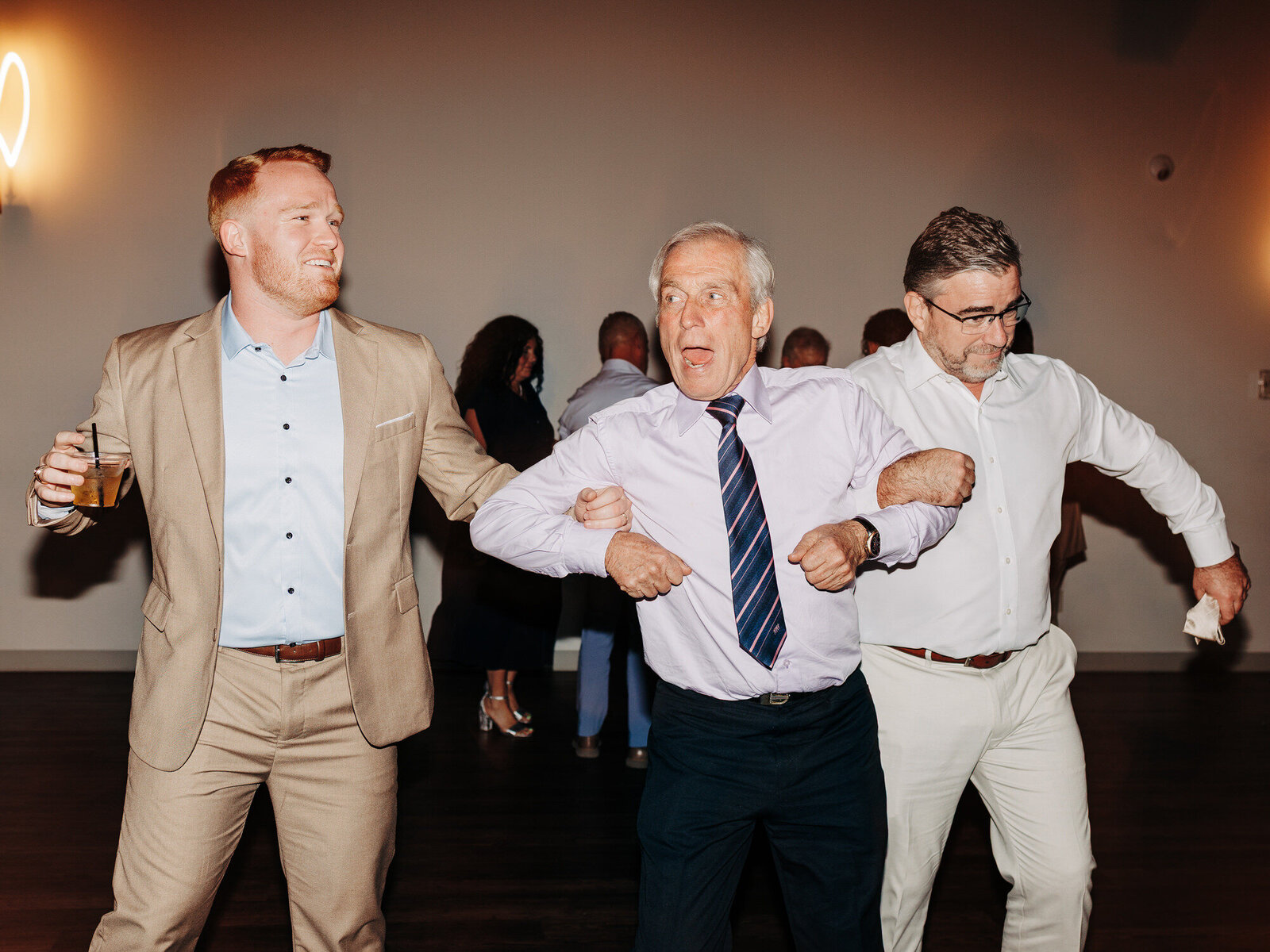 Three men, dressed in dress shirts and suits, link arms and dance enthusiastically at an indoor event. One man holds a drink, and all appear to be enjoying themselves. The background is softly lit with other guests visible, captured by an Austin wedding photographer.



