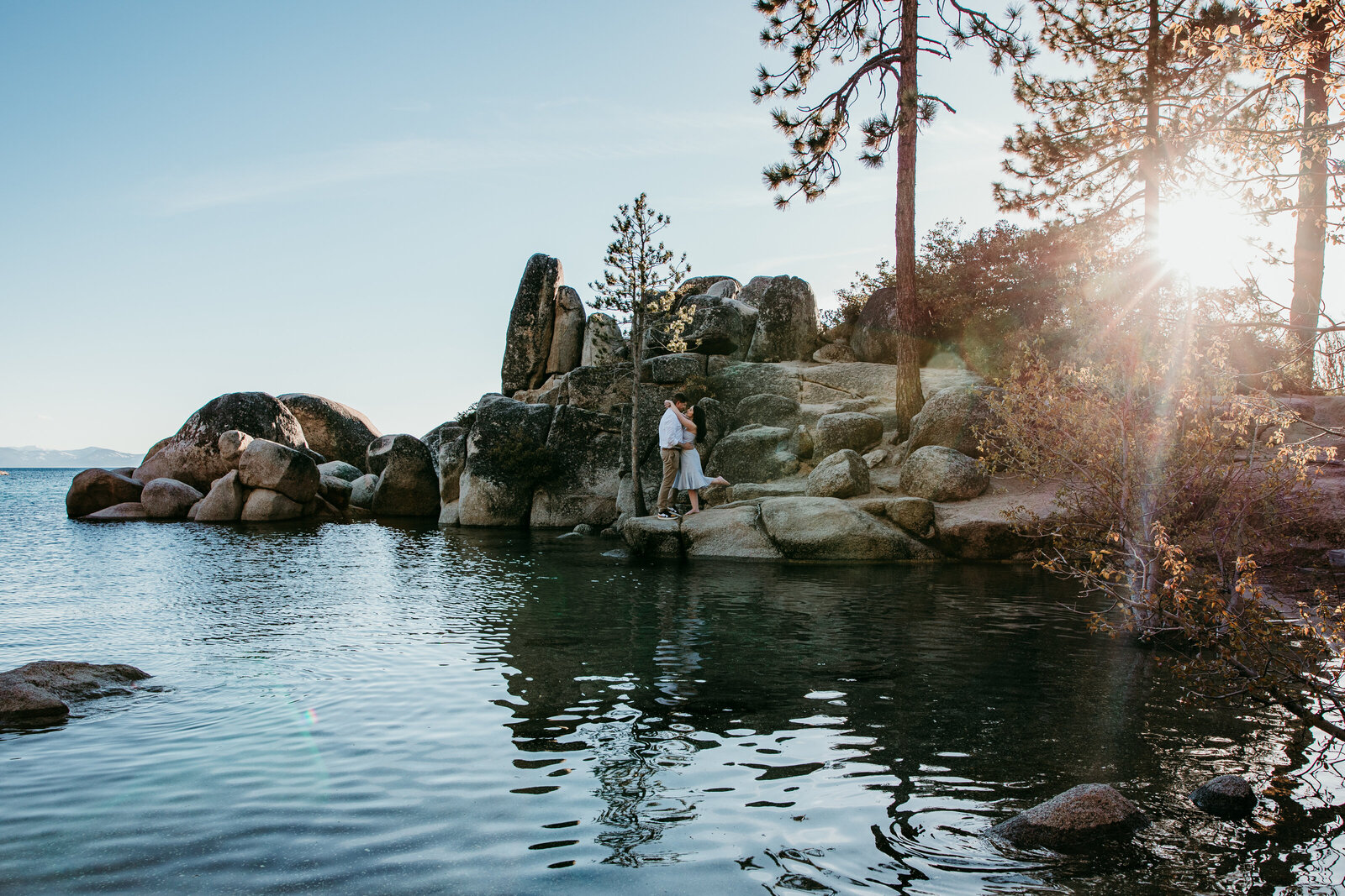 A couple embraces on the rocky shoreline of Lake Tahoe, surrounded by crystal-clear water and golden sunlight streaming through the trees.