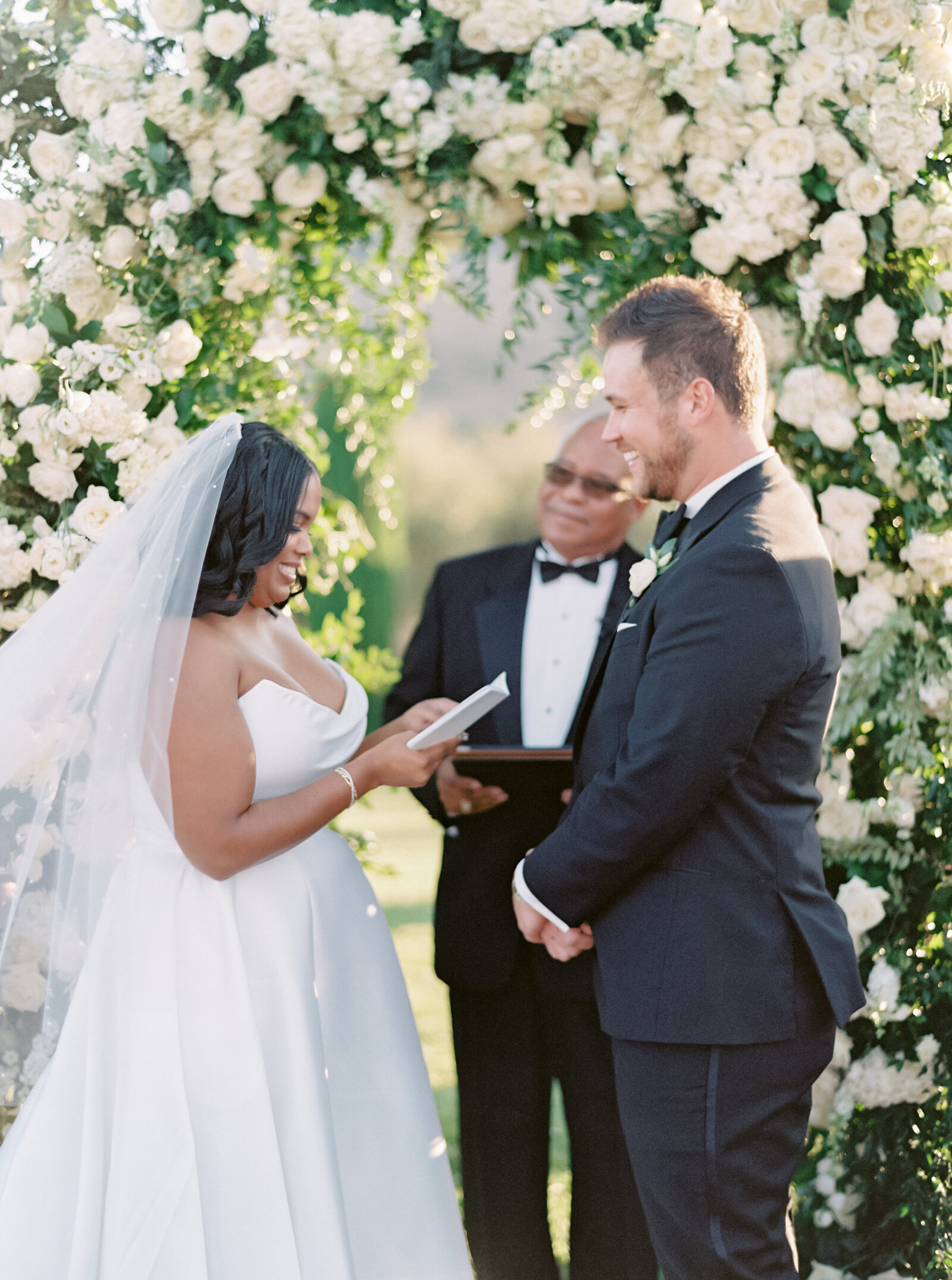 bride and groom exchanging vows under an arbor of white roses