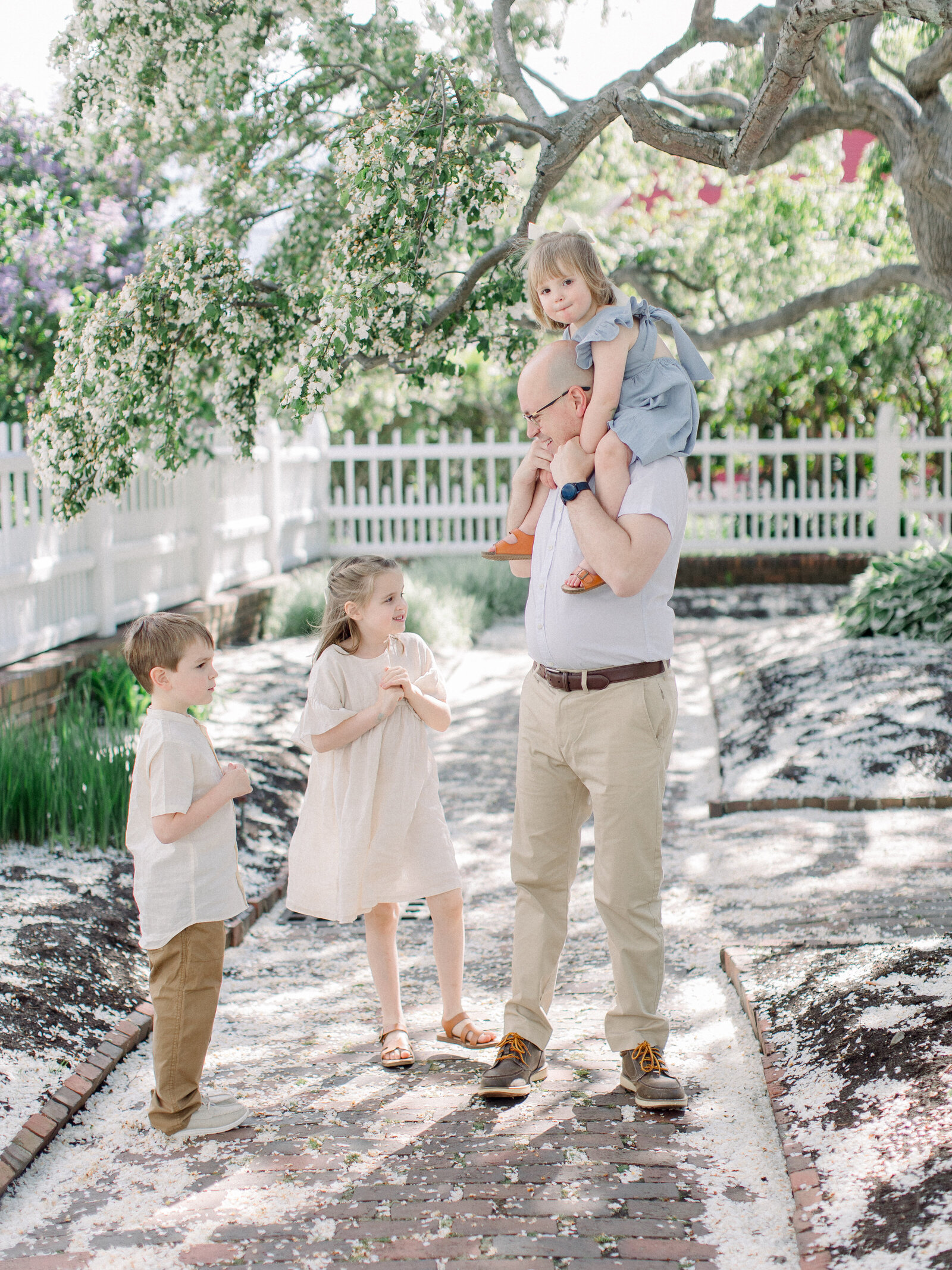 Dad with toddler sitting on his shoulders, standing with two older siblings in a spring garden under flowering cherry trees by NH newborn photographer Fieldstone Studio.