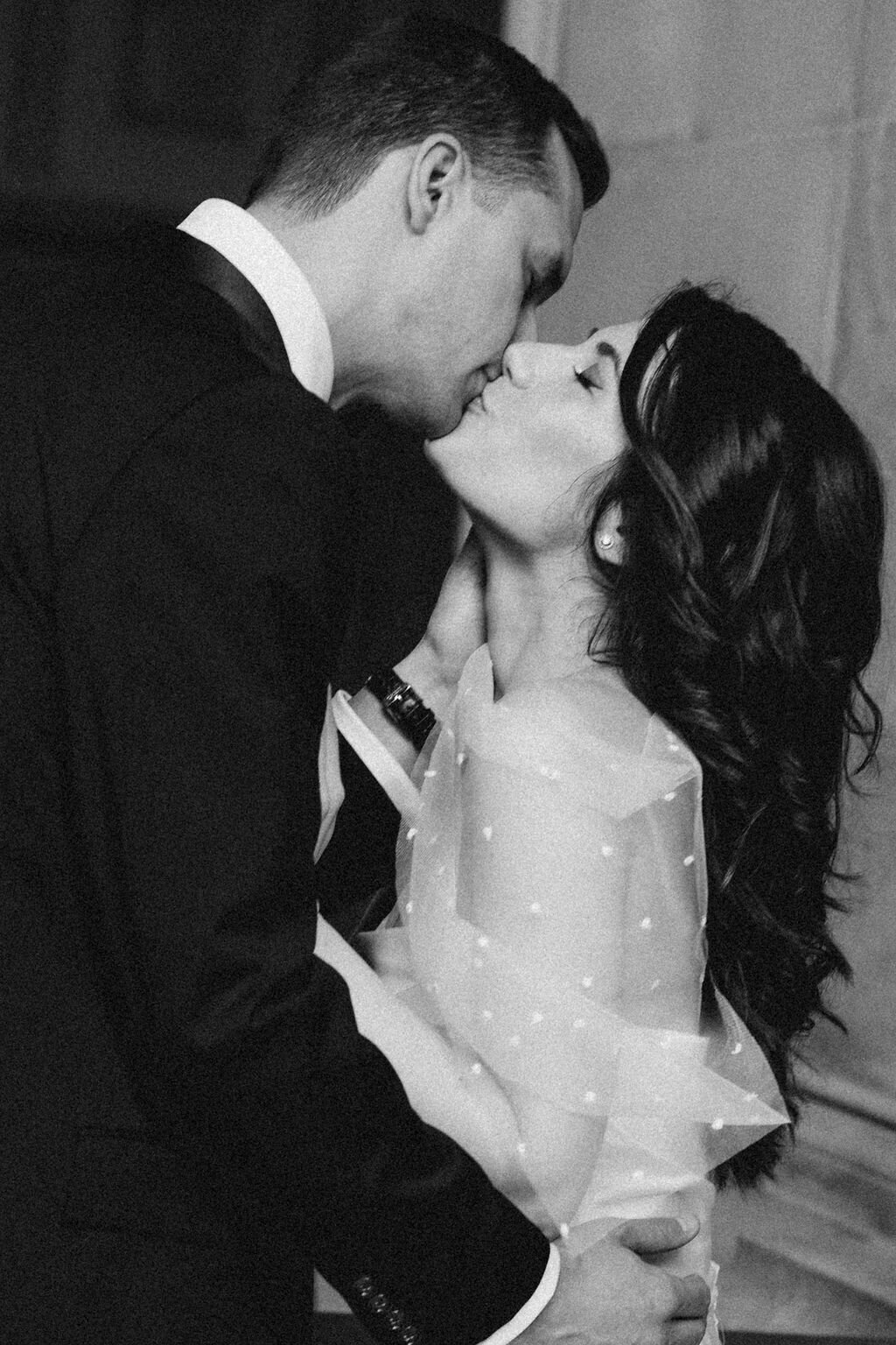 A monochrome image from a San Francisco City Hall photoshoot of a couple dressed in formal attire sharing a romantic kiss. The man is in a suit, and the woman in a dress with sheer polka-dot