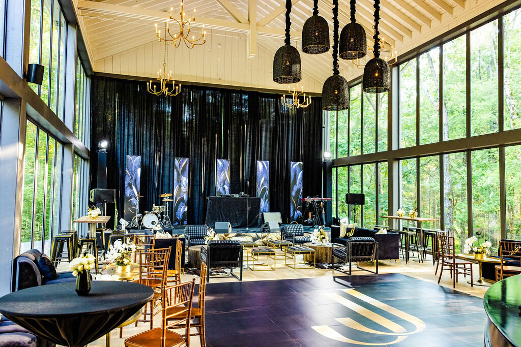 Chic black and gold event lounge at Mallard’s Croft featuring modern velvet seating, custom stage with drum kit, DJ booth, and keyboard, surrounded by floor-to-ceiling windows and forest views, styled by luxury event designer Wells Design Co.