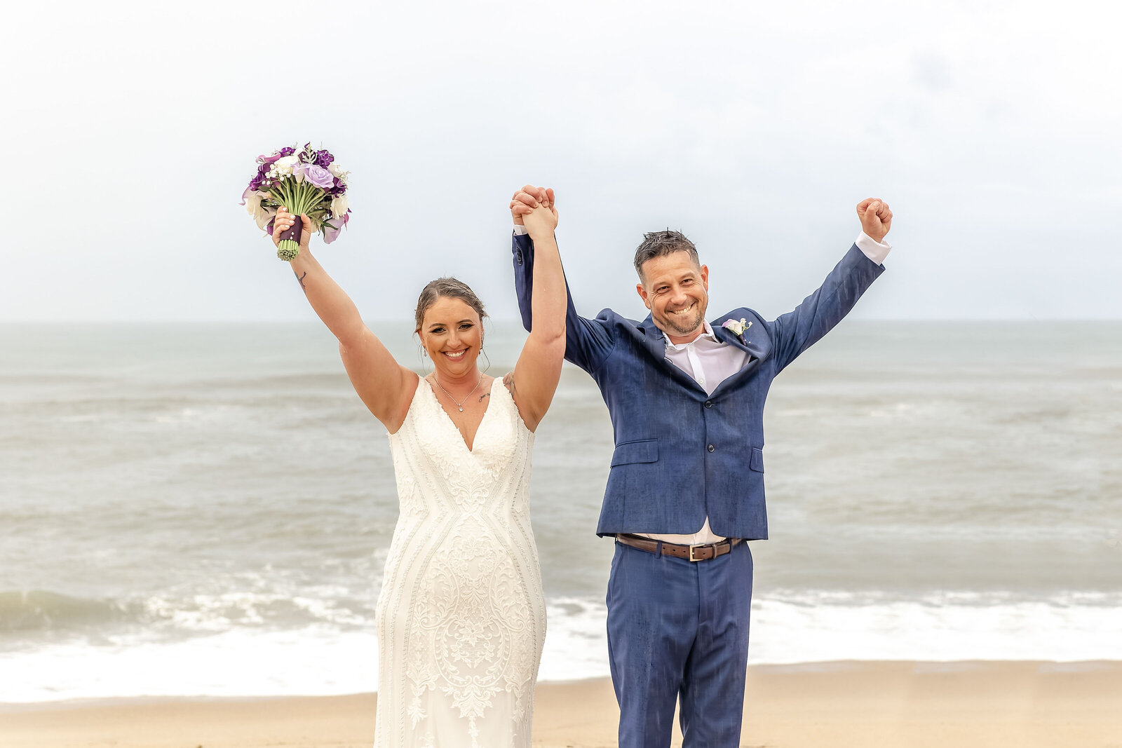 The bride and groom raise their hands in celebration on the sandy shores of Outer Banks, NC, with soft waves in the background, showcasing coastal charm and joyful wedding moments.