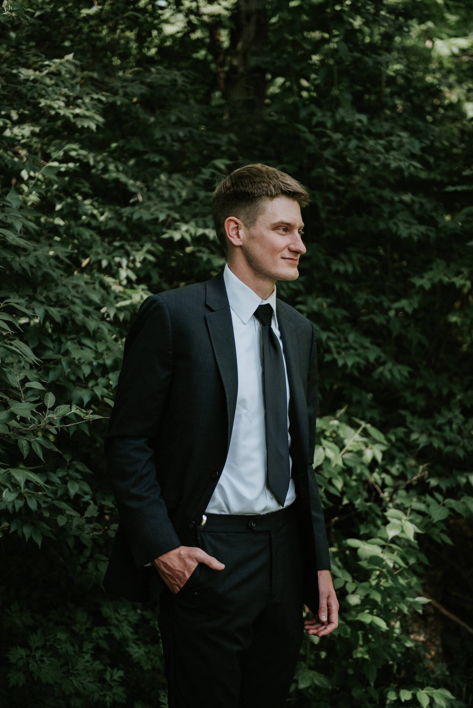 A groom stands with one hand in his pocket in the woods