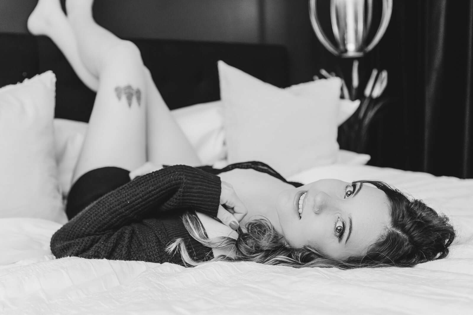 A black-and-white photo of a woman relaxing on a bed, smiling, with her legs raised playfully in the air.