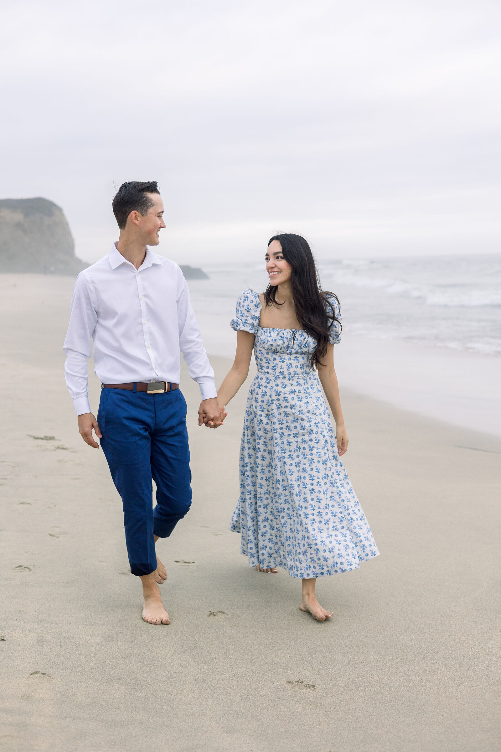 wedding photographer bay area captures proposal pictures at half moon bay while walking on the beach together and smiling as they hold hands