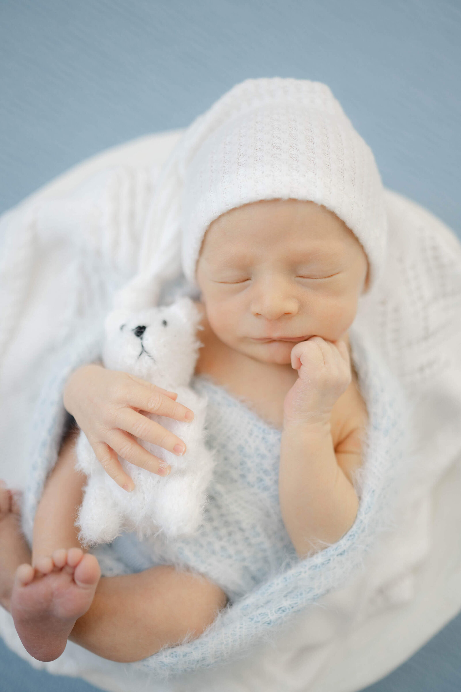 adorable baby swaddled in blue and wearing a sleepy hat cuddling a fuzzy mini teddy bear