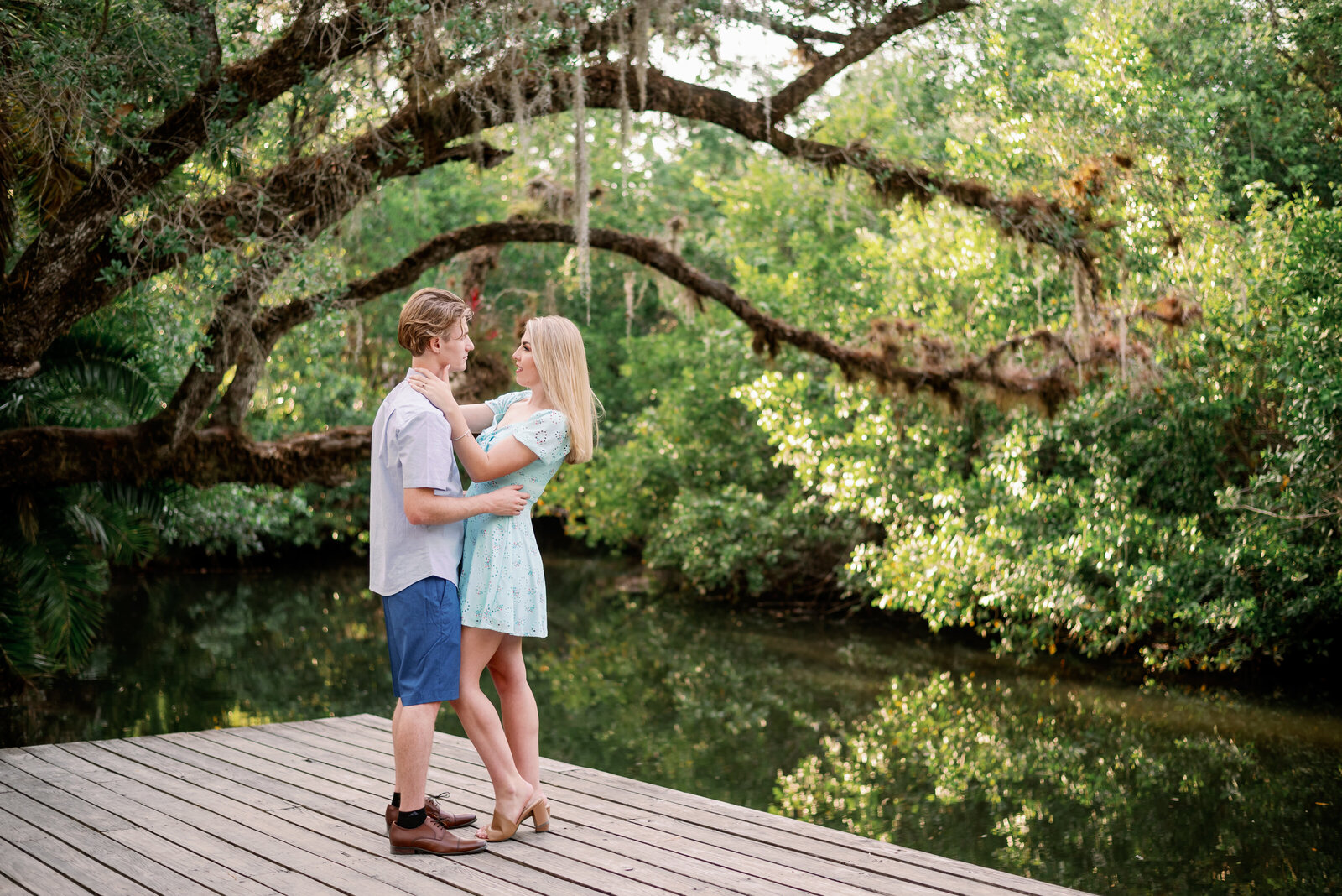 wide view photograph of engaged couple standing on a dock, looking at each other. The Estero  River and old oak trees are behind them