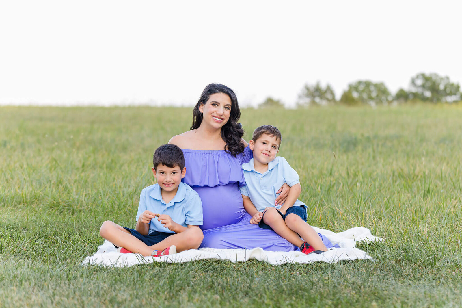 A mom-to-be sitting in the grass with her older two boys.