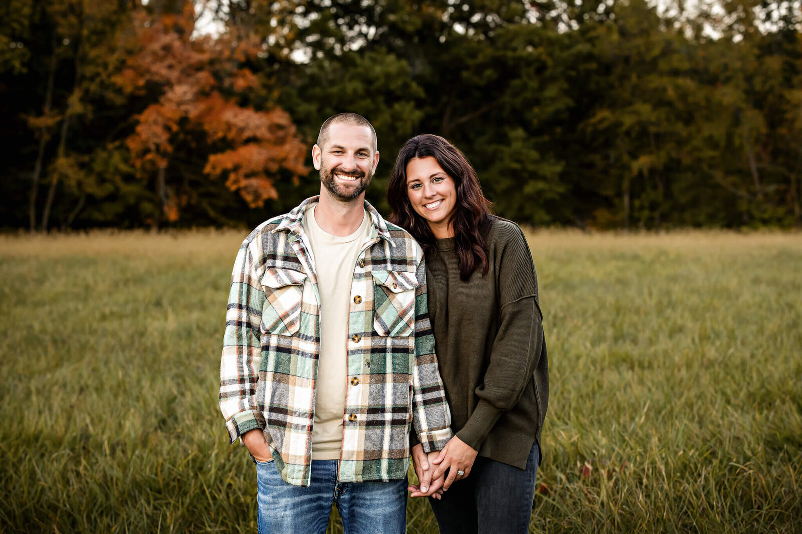 A smiling couple stands in a grassy field, captured beautifully in outdoor portraits. The man wears a plaid jacket and jeans, while the woman is in a green sweater and jeans. They hold hands amidst trees and fall foliage—perfectly framed by a talented Bloomington family photographer.