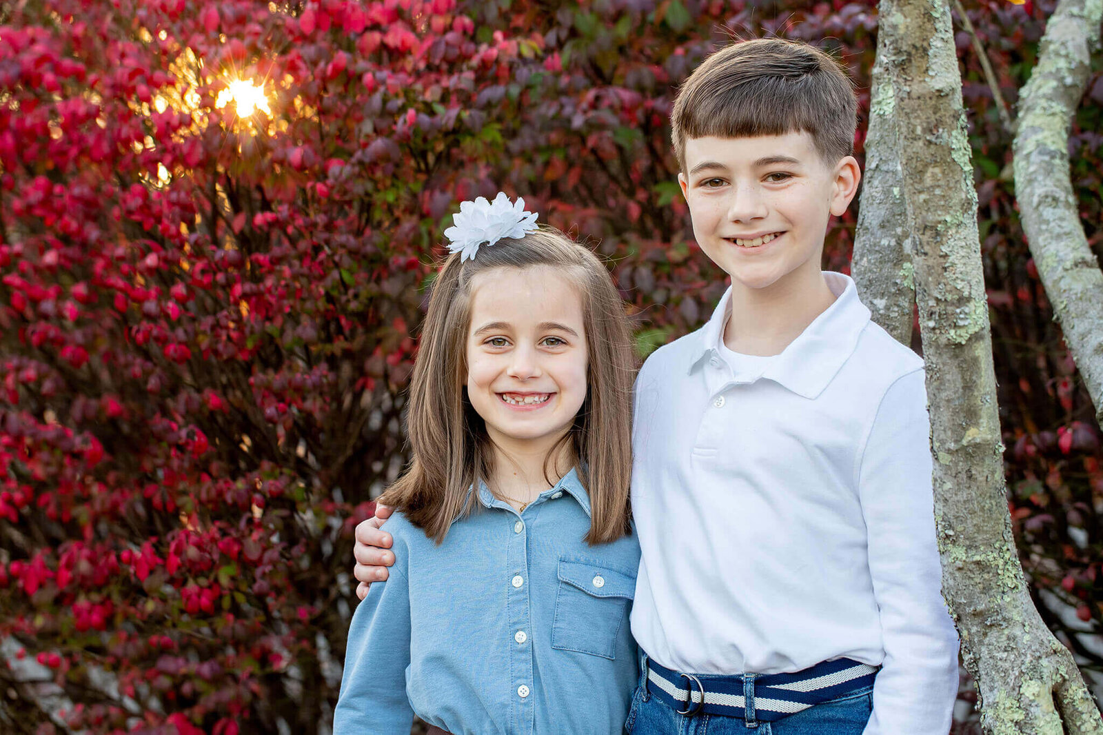 A brother and sister standing in front of a red bush with the sun shining through.