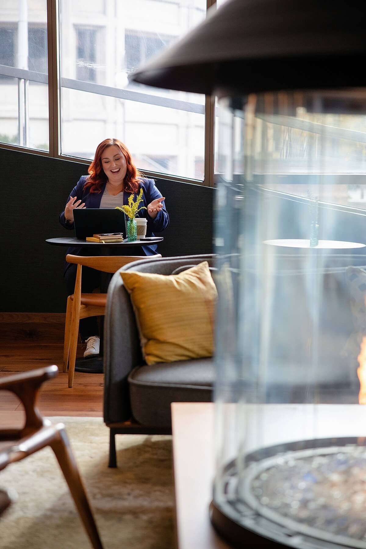 Woman entrepreneur laughing during a branding photoshoot in a modern Seattle workspace, captured by a Seattle brand photographer. She is seated at a cozy café-style table with her laptop, embodying confidence and connection. Personal branding photography session for “She Plays With Fire,” showcasing storytelling-driven visuals for business owners.