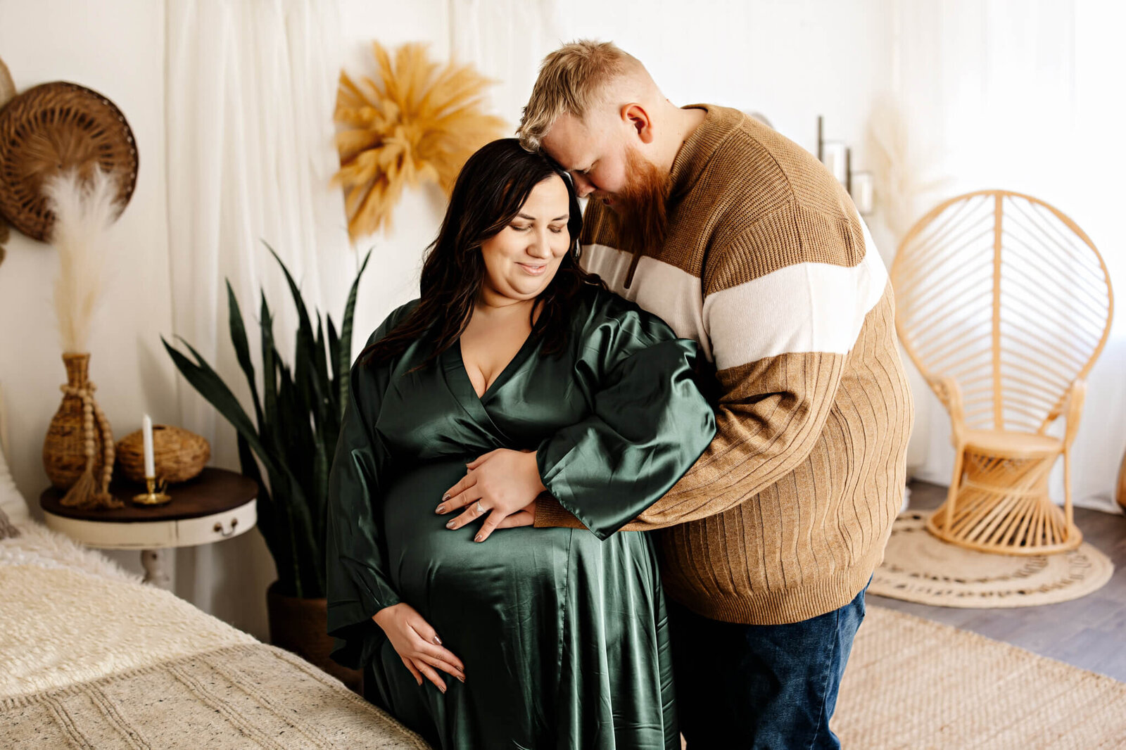 A pregnant woman in a green dress stands indoors, smiling as a man in a brown sweater hugs her from behind and gently rests his hand on her baby bump—a perfect moment for maternity portraits surrounded by plants and warm-toned decor.