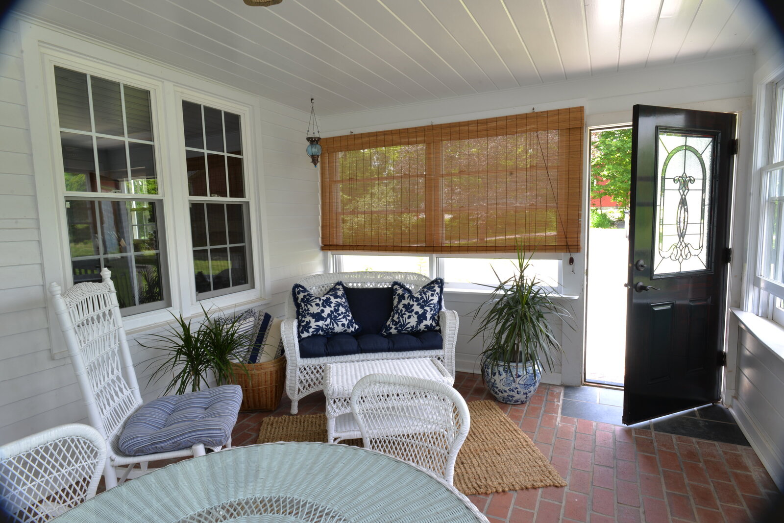 sunroom of farmer cottage with white walls and brick floor and woven roman shades with white wicker seating