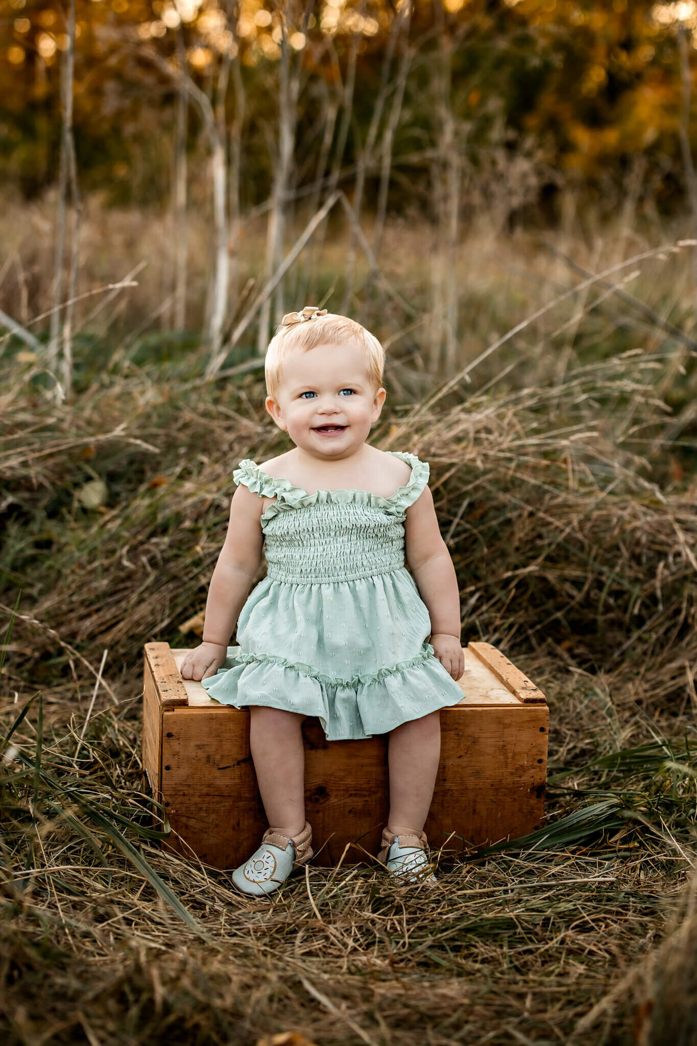 A smiling toddler sits on a wooden box in a grassy field, showcasing the magic of outdoor portraits. She is wearing a light green dress and matching shoes as golden light from the setting sun illuminates the tall grass and trees in the background.