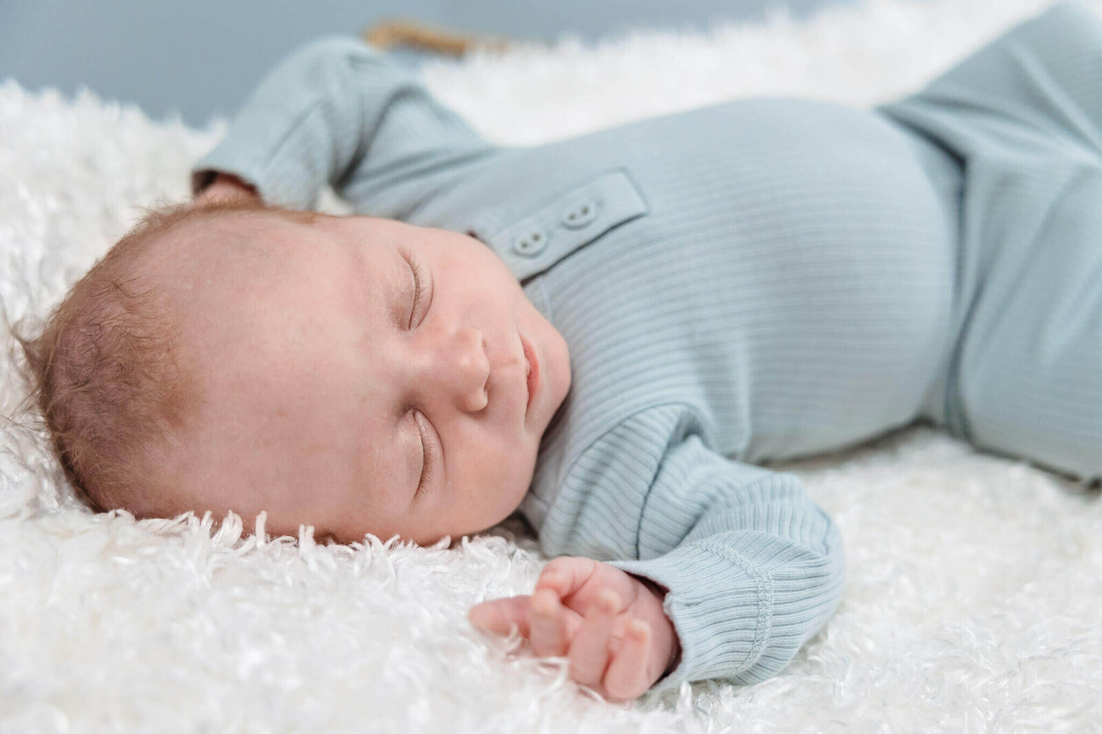 A newborn boy in blue asleep in a Moses basket filled with a fluffy white blanket.