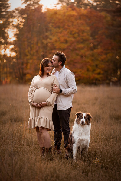 Een aanstaande moeder en haar partner poseren liefdevol met hun hond tijdens een zwangerschapsshoot, een intiem moment vol warmte en verwachting