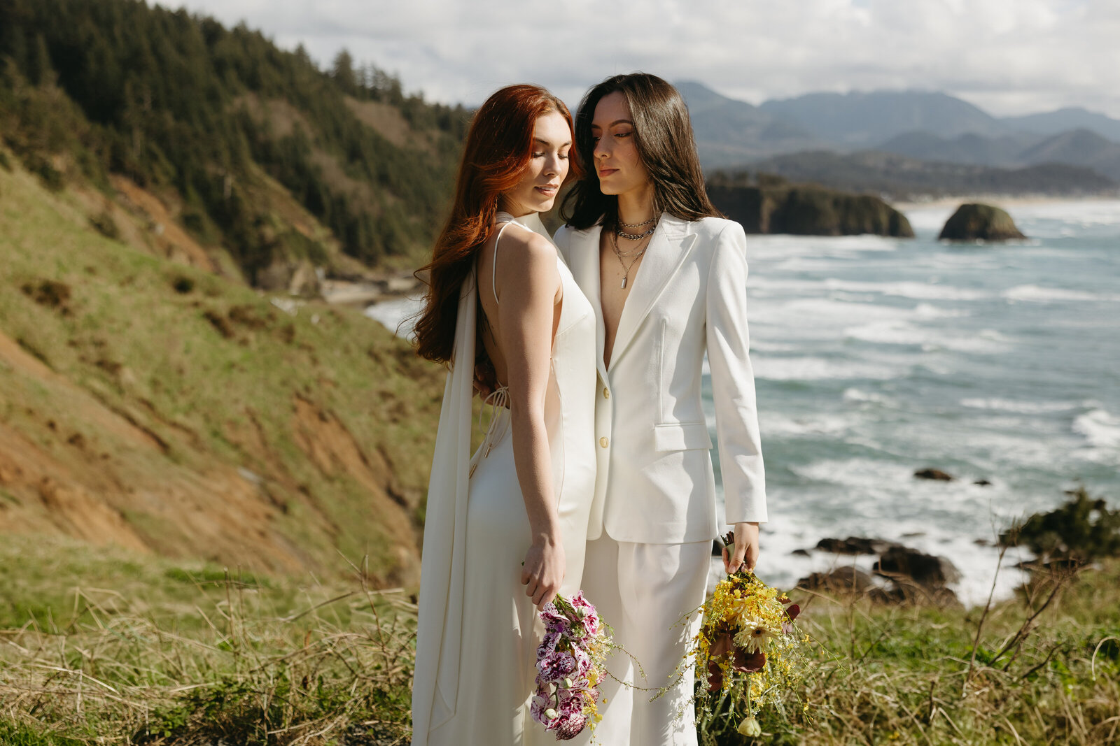 Two brides stand together along the coast of the Pacific ocean after eloping. 
