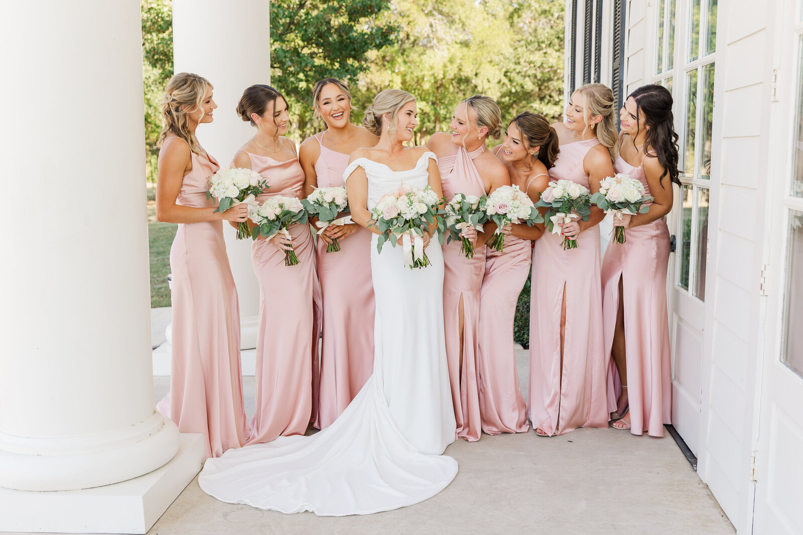 Bride in an off-the-shoulder gown surrounded by bridesmaids in blush pink satin dresses, all holding soft white and blush rose bouquets, smiling and laughing outside a white-columned venue in Denton, Texas.