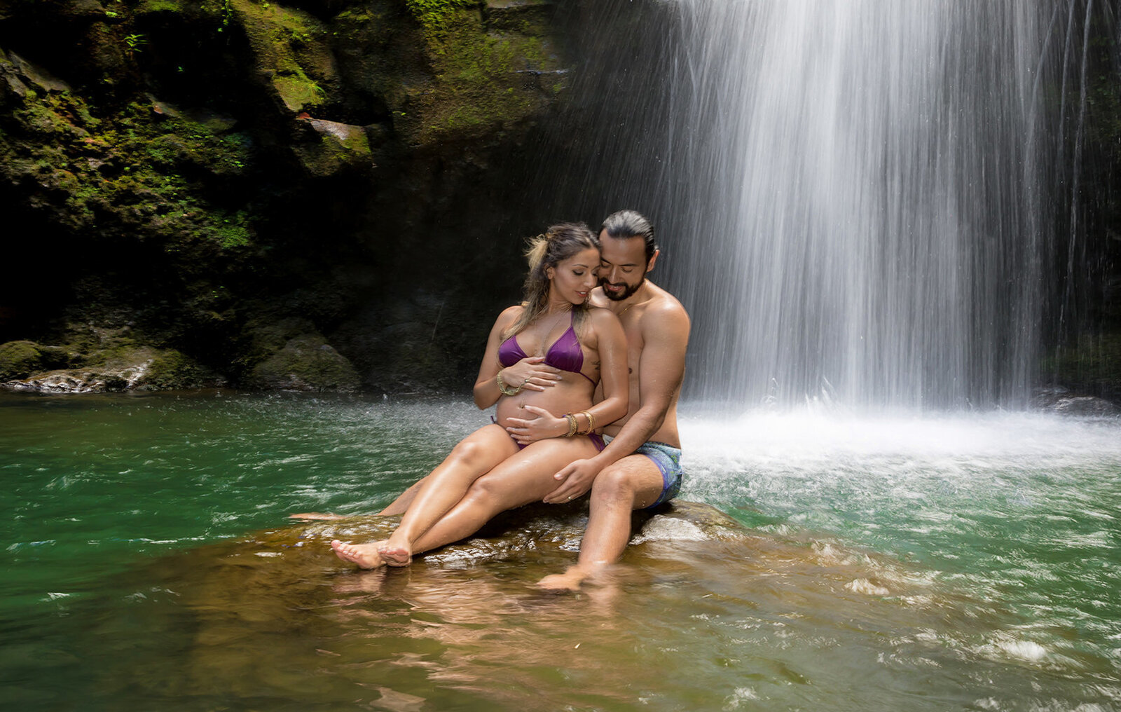 Couple sitting on a rock where a  man has arms around his wives pregnant belly surrounded by a big waterfall with water running all around them in Oahu