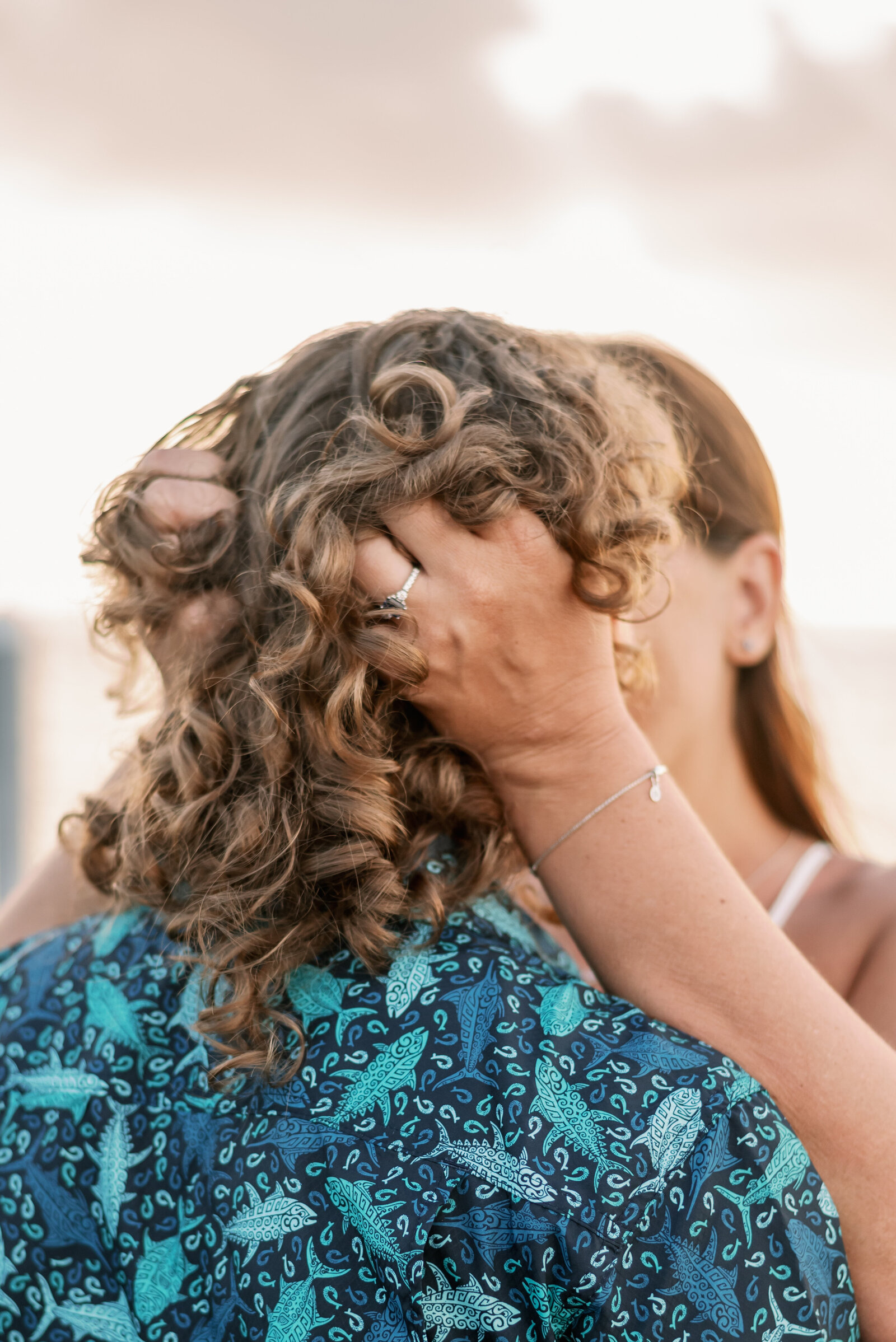 LGBTQ engaged couple kissing, we see one womans hands showing her engagement ring with her fingers entwined into the back of the other womans hair