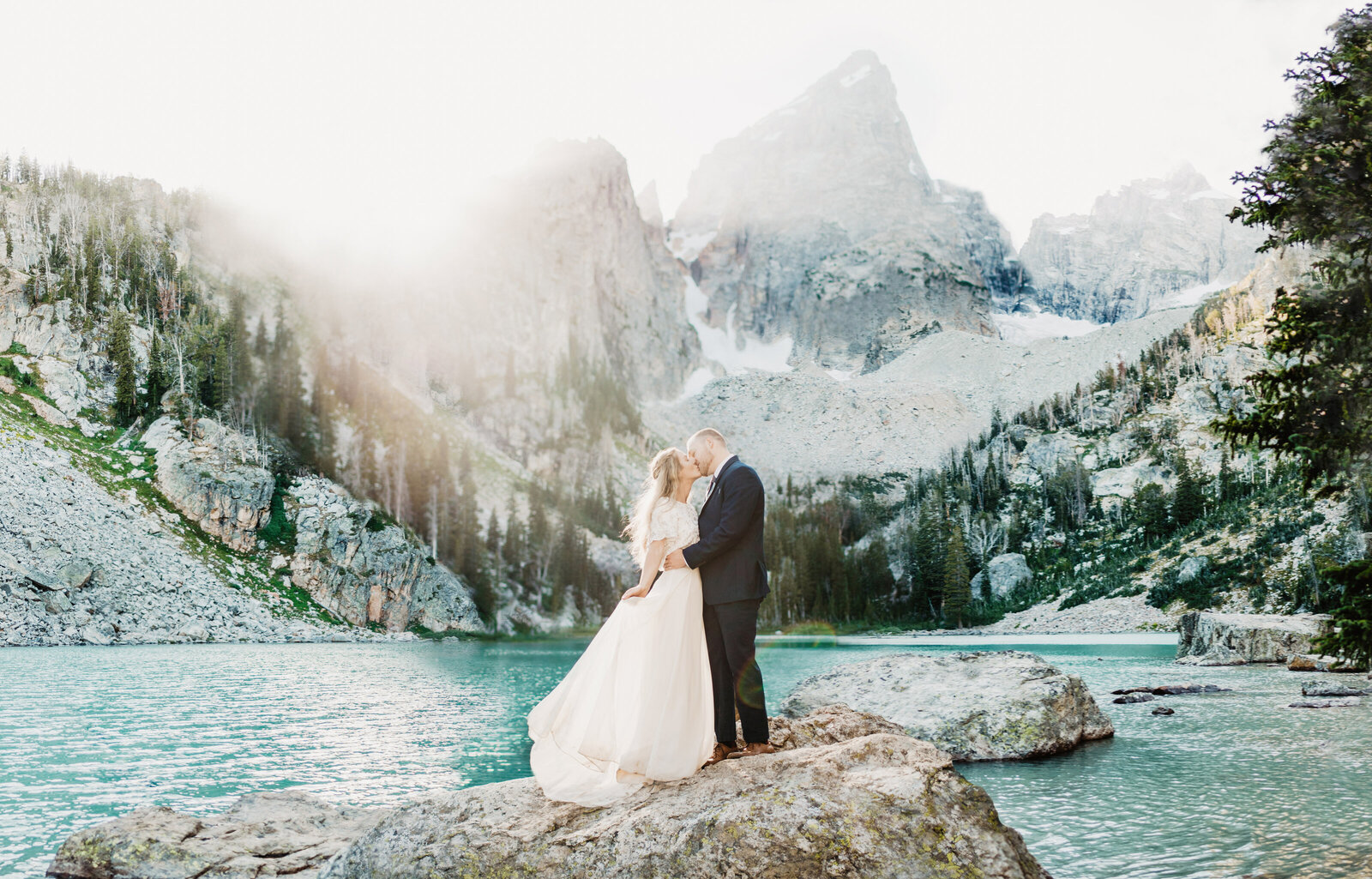 wyoming elopement with bride and groom standing on a rock in the middle of Delta Lake Wyoming  with the sun shining through the mountains captured by jackson hole photographers