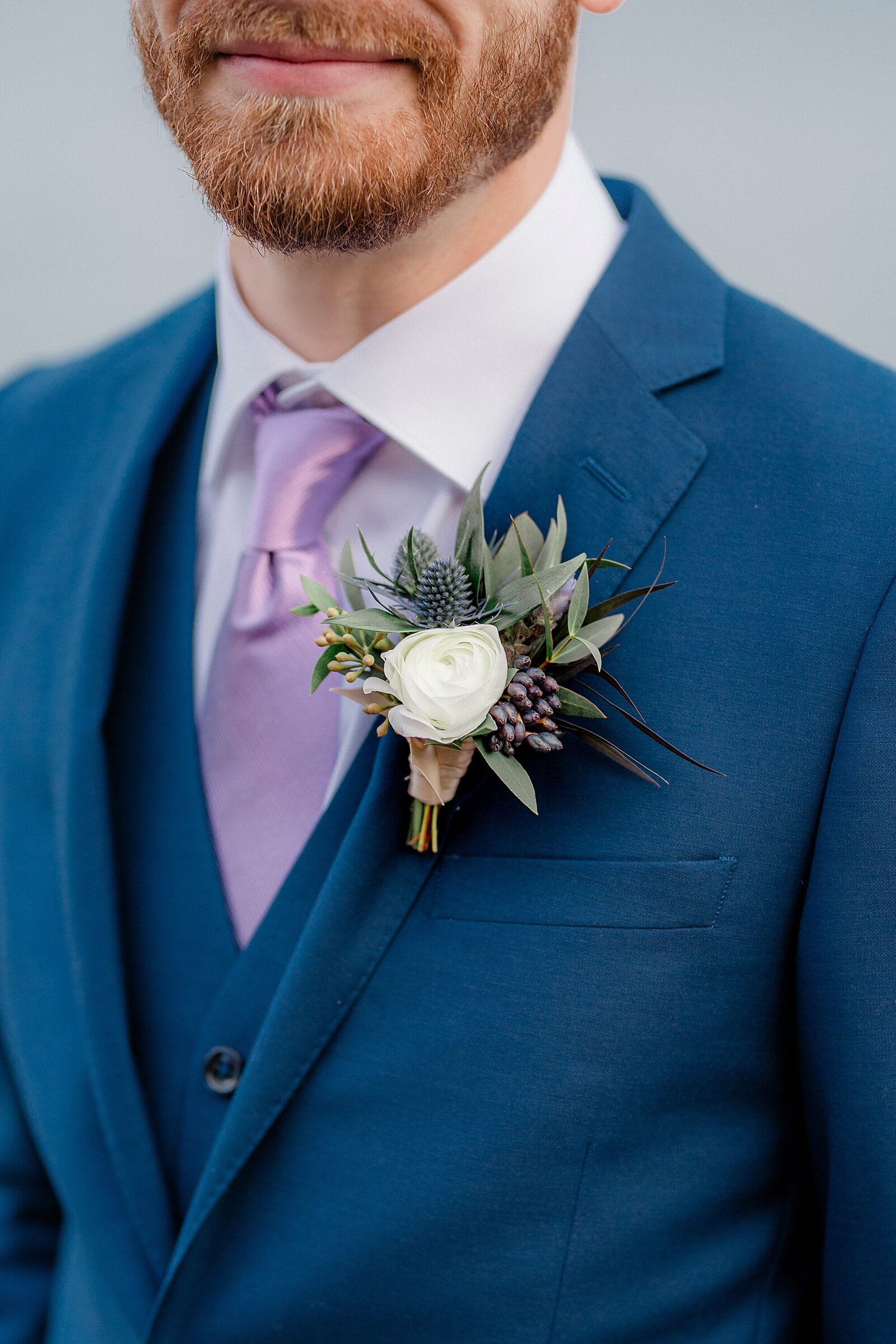 This is a close up of a boutonniere, which features a while rose. The man is wearing a lilac tie an blue suit, which is what he wore to his Minnesota wedding.