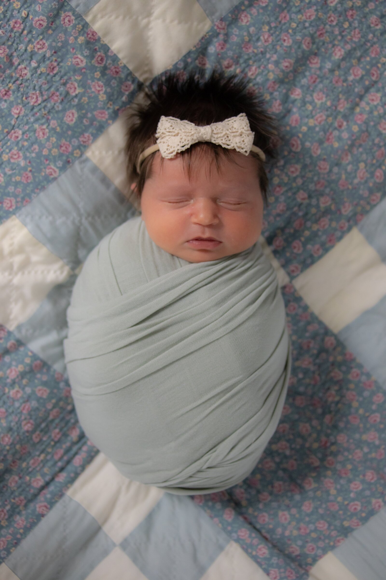 Swaddled newborn baby girl in a sage green wrap and bow on top of dad's baby blanket.