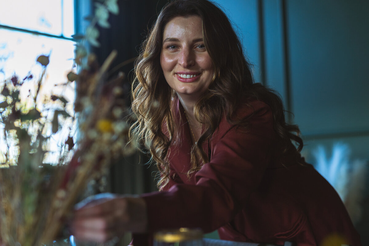 A smiling woman with long brown hair arranges flowers indoors, reflecting her expertise as a luxury event planner in Northwest Arkansas.