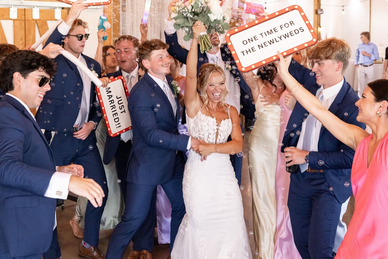 A joyful wedding celebration as the newlywed couple makes their grand entrance into the reception. The bride, beaming with excitement, raises her bouquet high in the air while the groom wraps his arms around her from behind. Friends and wedding party members surround them in a tunnel of cheering, waving foam sticks and holding playful signs that read “JUST MARRIED!!” and “ONE TIME FOR THE NEWLYWEDS.” Everyone is dressed in elegant formalwear—men in navy suits and women in satin dresses—and the space is filled with bubbles, laughter, and high energy.