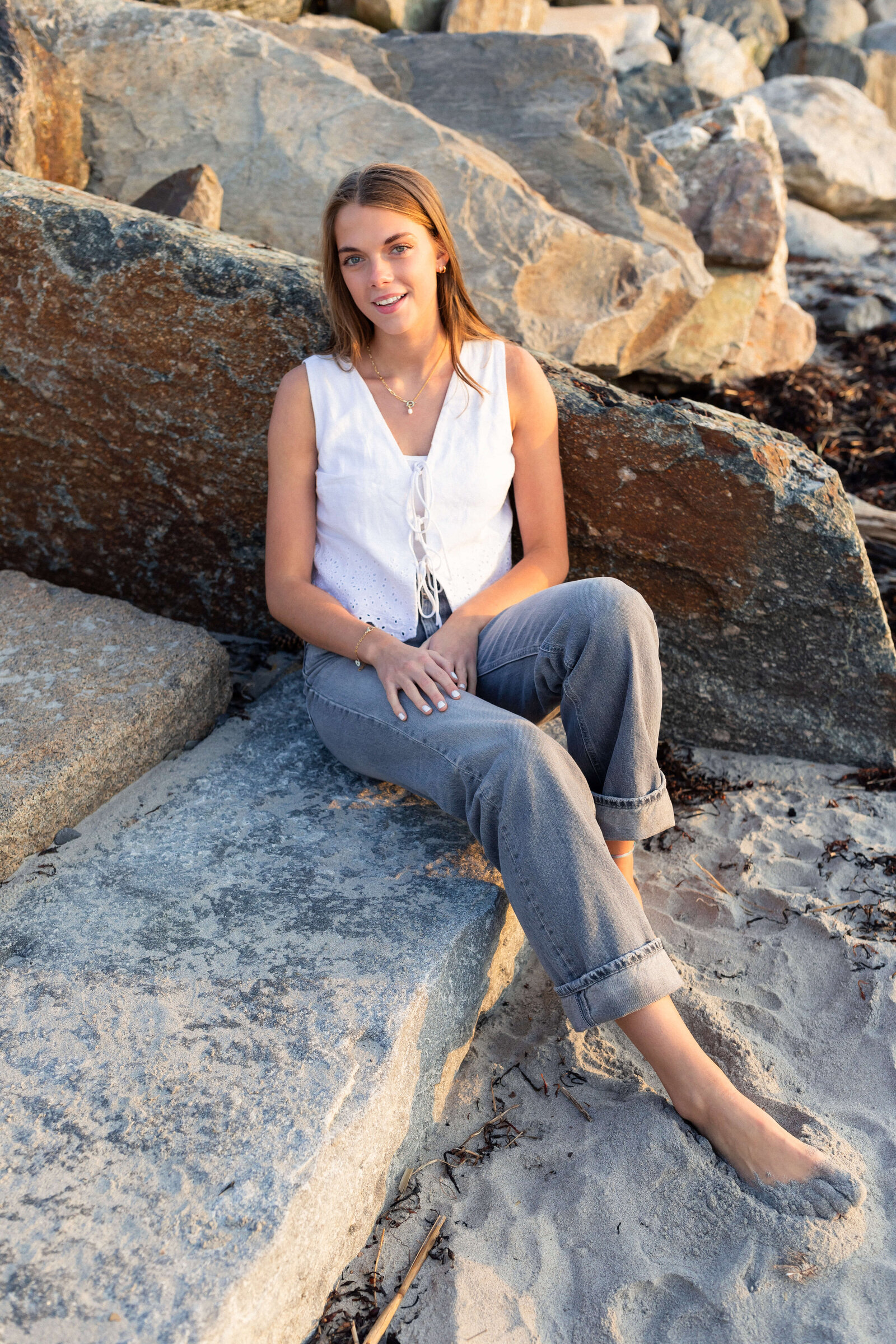 brunette high schooler posing on a beach wearing a white shirt and blue jeans on a sandy, rocky beach