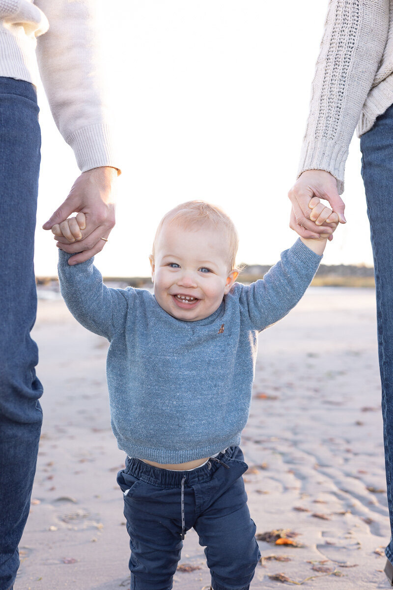 Smiling toddler holding hands with parents during a joyful lifestyle family session on the beach in Westchester County. Captured by Nicole, a family photographer serving Fairfield and Westchester families who love natural, heartwarming photos.