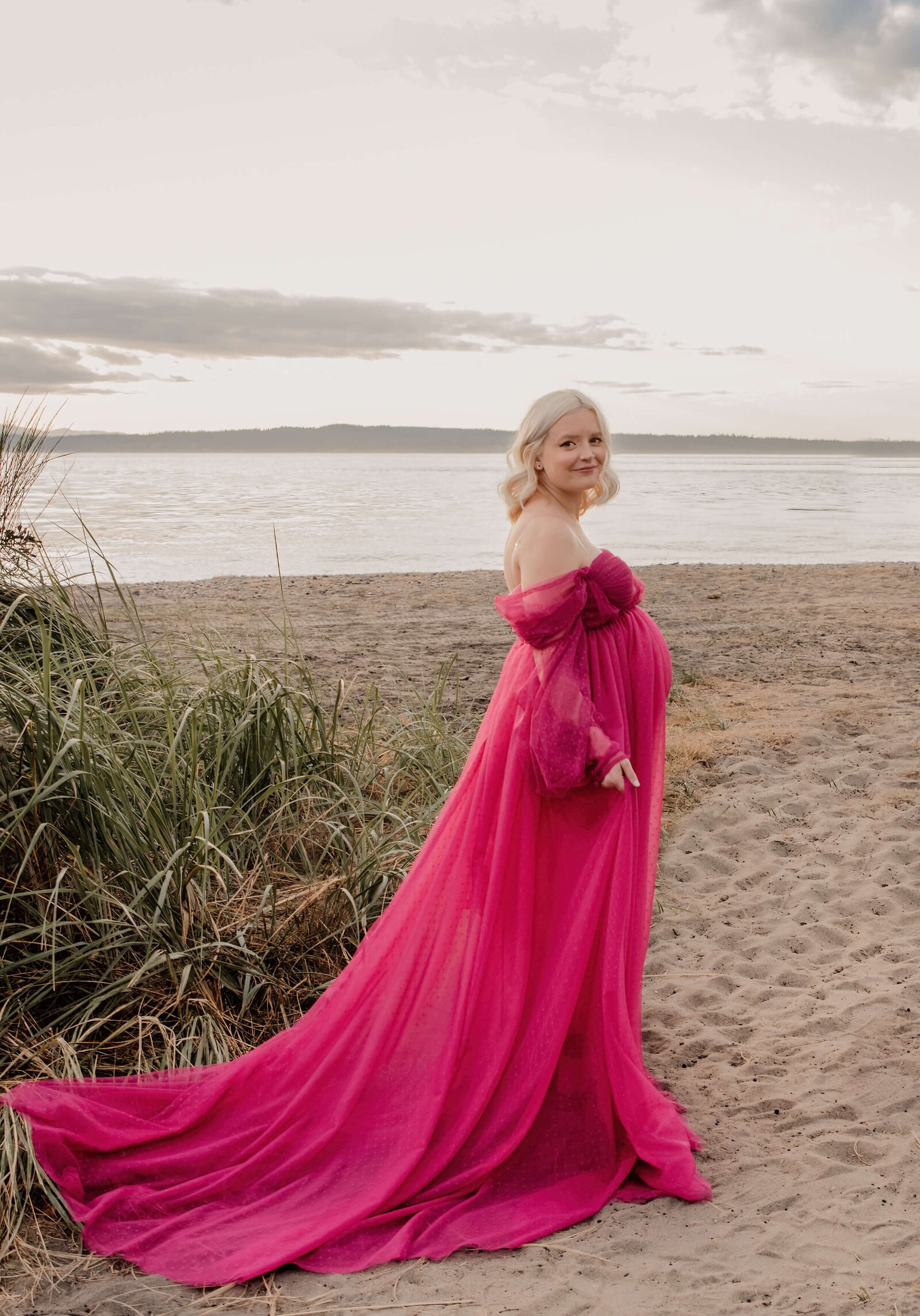 Pregnant woman in pink dress standing on beach.