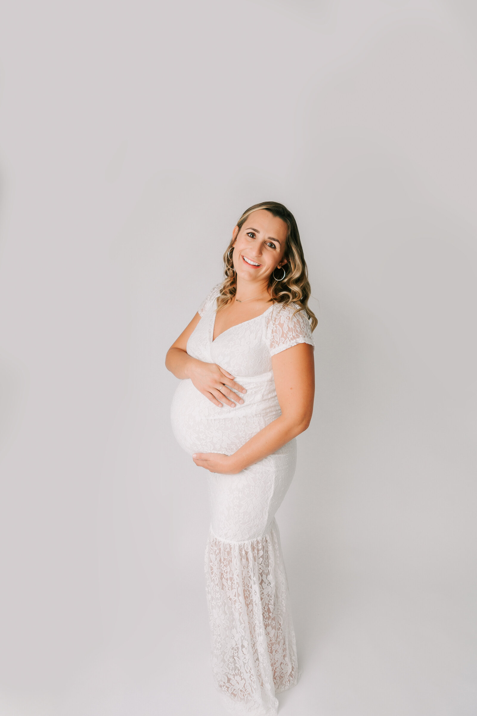 Studion maternity photography. Woman is photographed wearing a white lace dress that shows off her beautiful pregnant belly