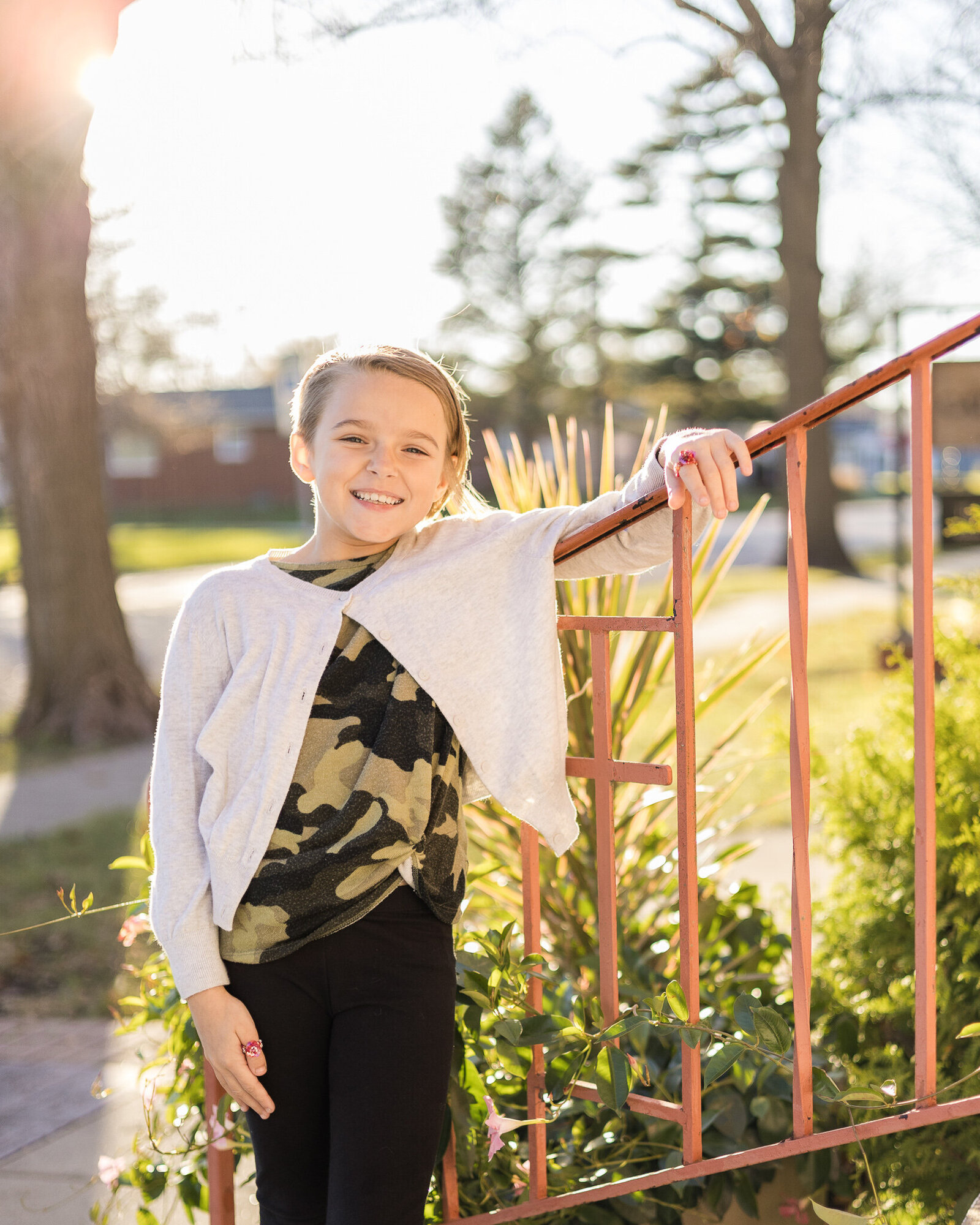 A young girl against a railing, with short sandy blonde hair, wearing a white button up sweater, camo shirt, and black pants, smiling at the camera