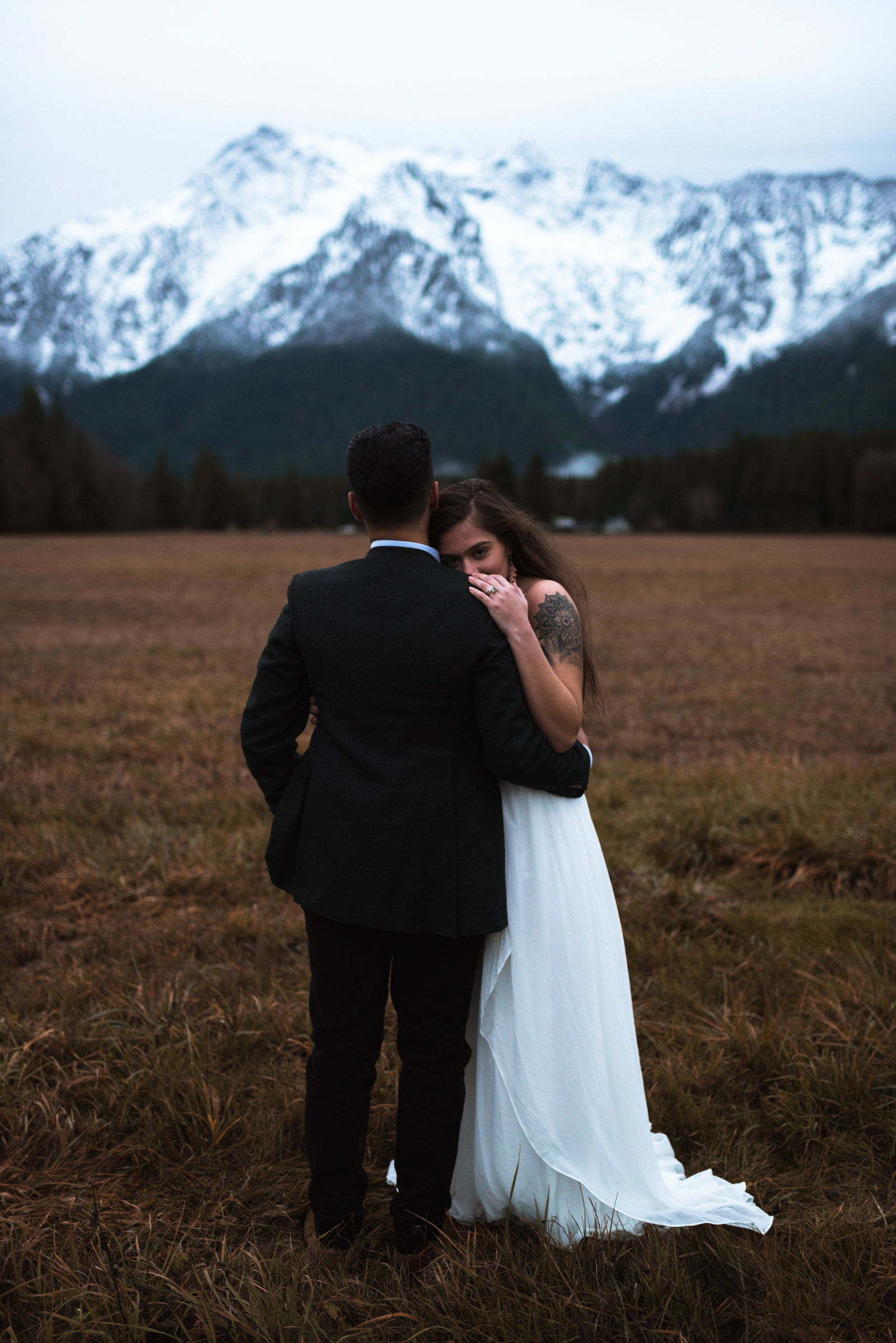 Couple in wedding attire, holding each other with snowy mountain in background.