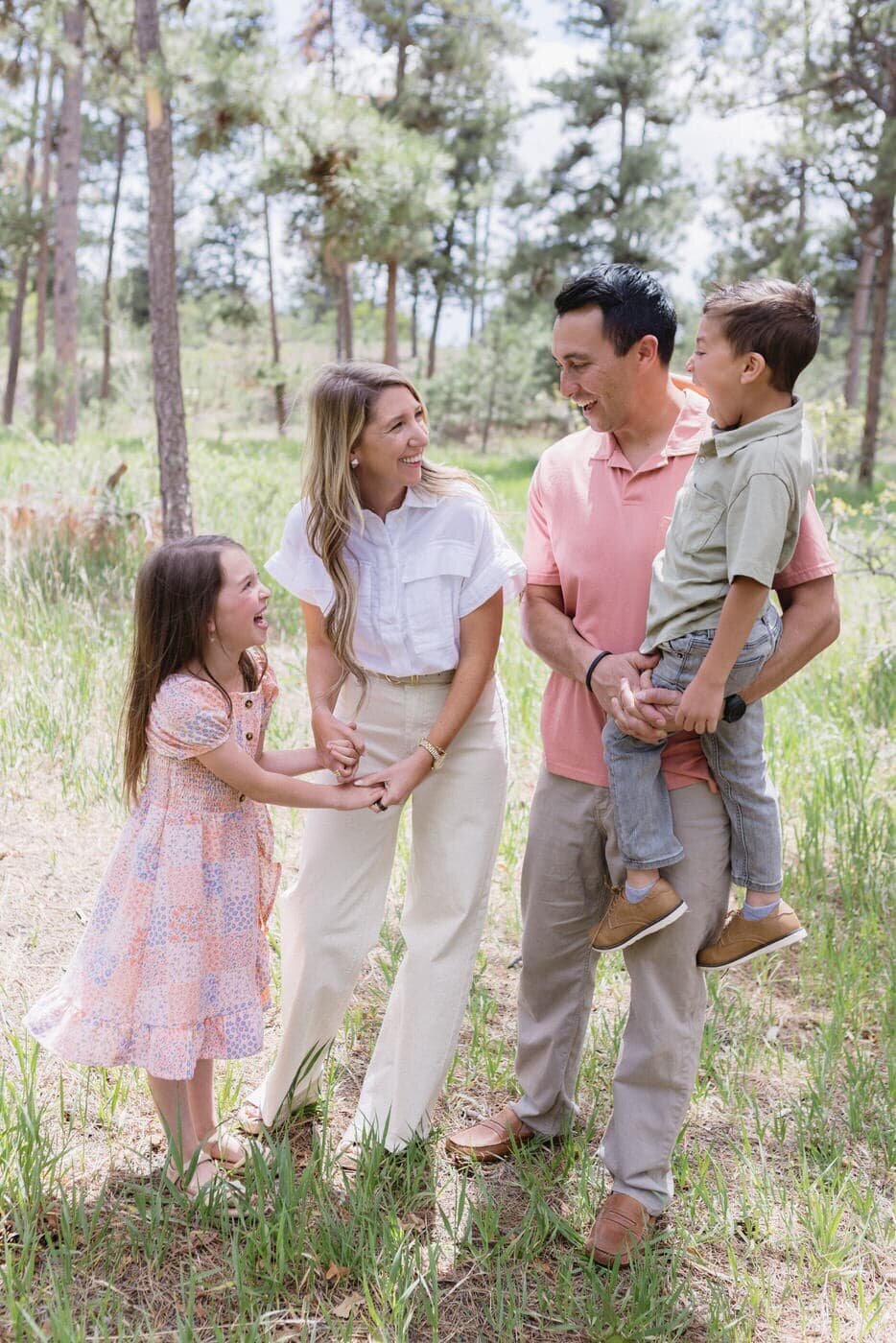 A family of four stands looking at each other and laughing in tall green grass with tall trees behind them.