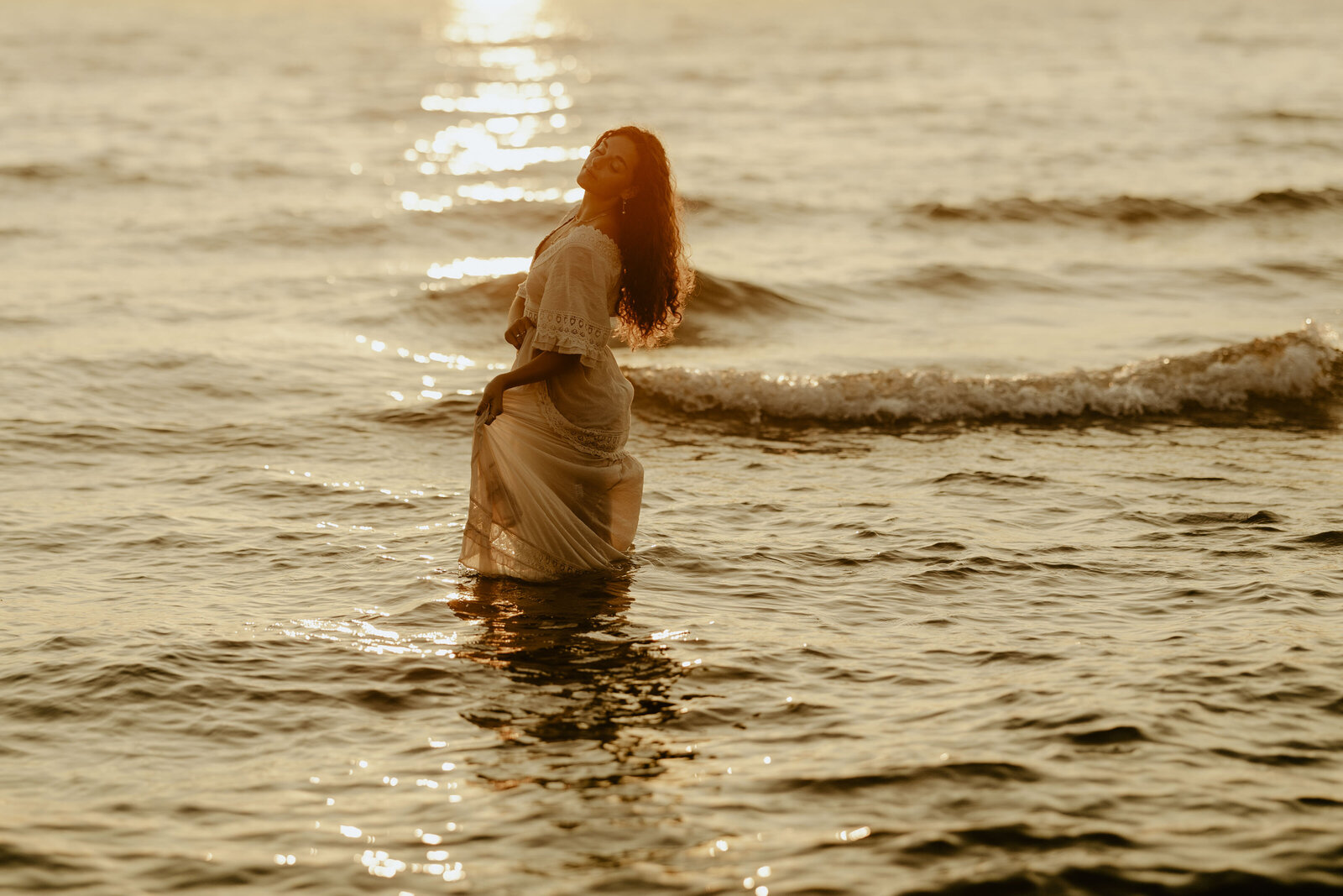 A girl holding her dress and closing her eyes while she stands in Lake Michigan at sunset