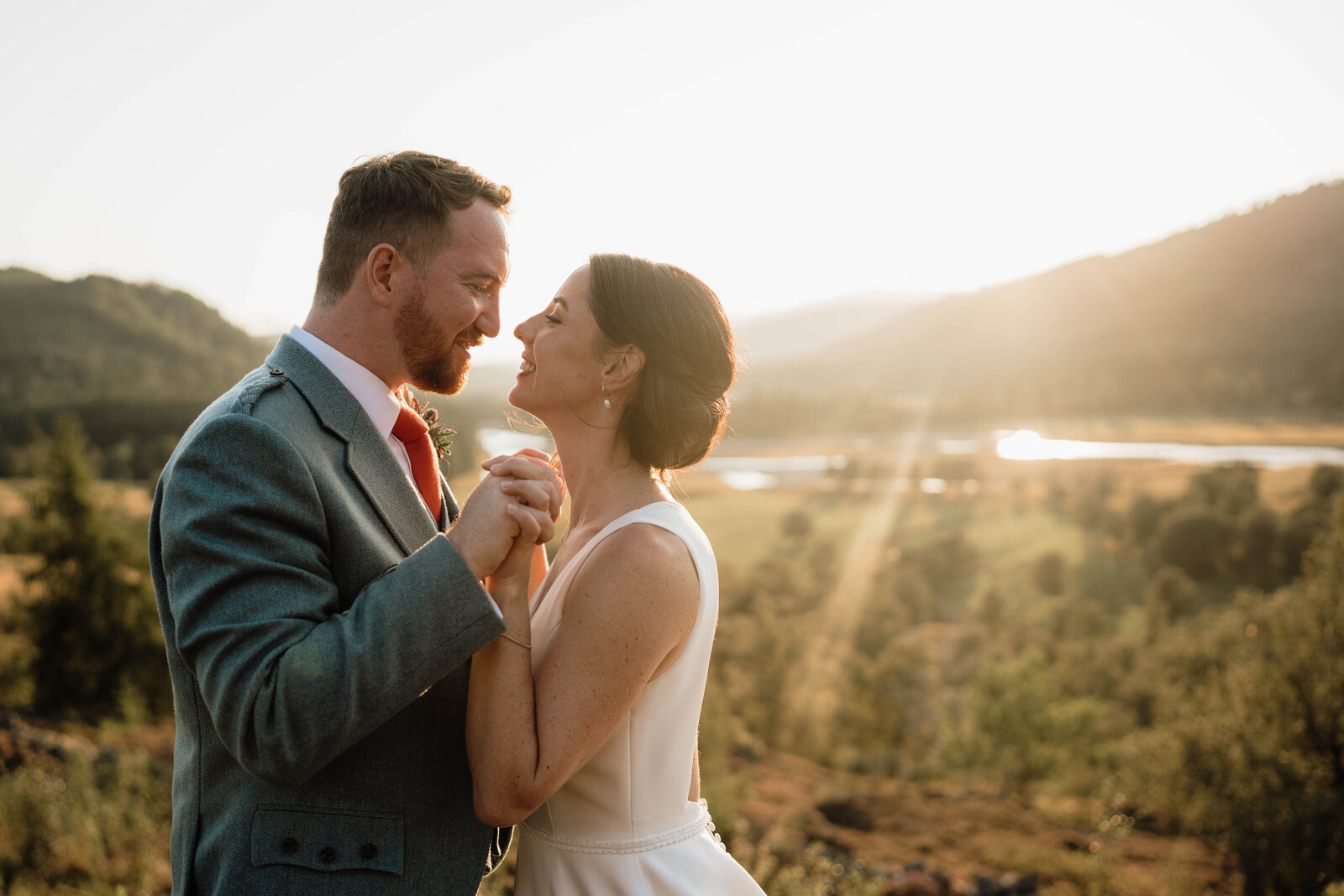 A close photography of a bride and groom linking fingers and touching chests. They stand in the Mar Lodge valley after heir wedding as the sun starts to set. The vallery glows behind them and they go to kiss.