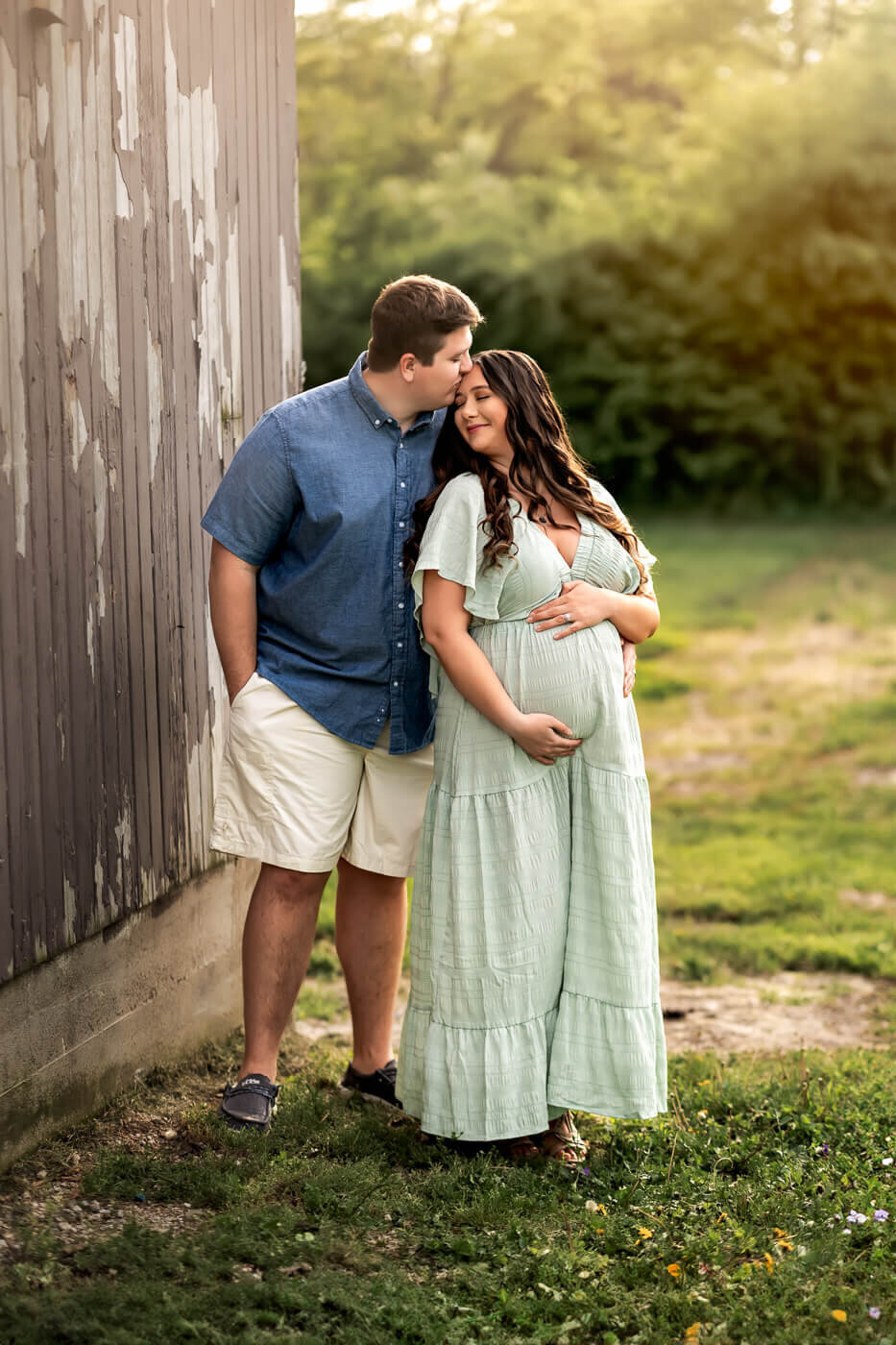 A couple stands outdoors by a weathered wooden wall. The man in a blue shirt gently kisses the forehead of his partner in a light green dress, lovingly cradling her belly—an intimate moment perfect for maternity portraits by a Bloomington maternity photographer.