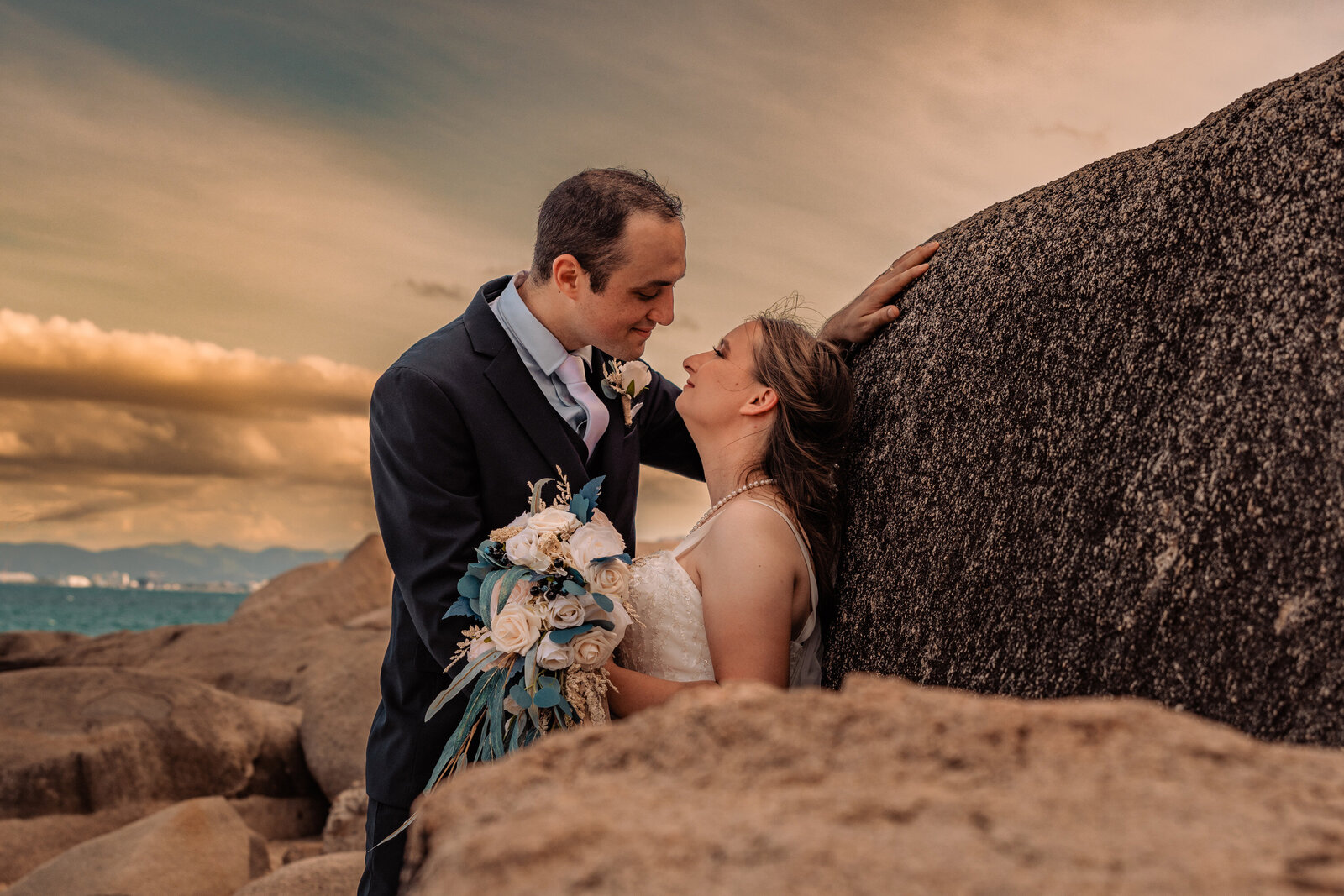 A couple in wedding attire embraces on rocky cliffs, surrounded by the vibrant colors of a sunset sky.