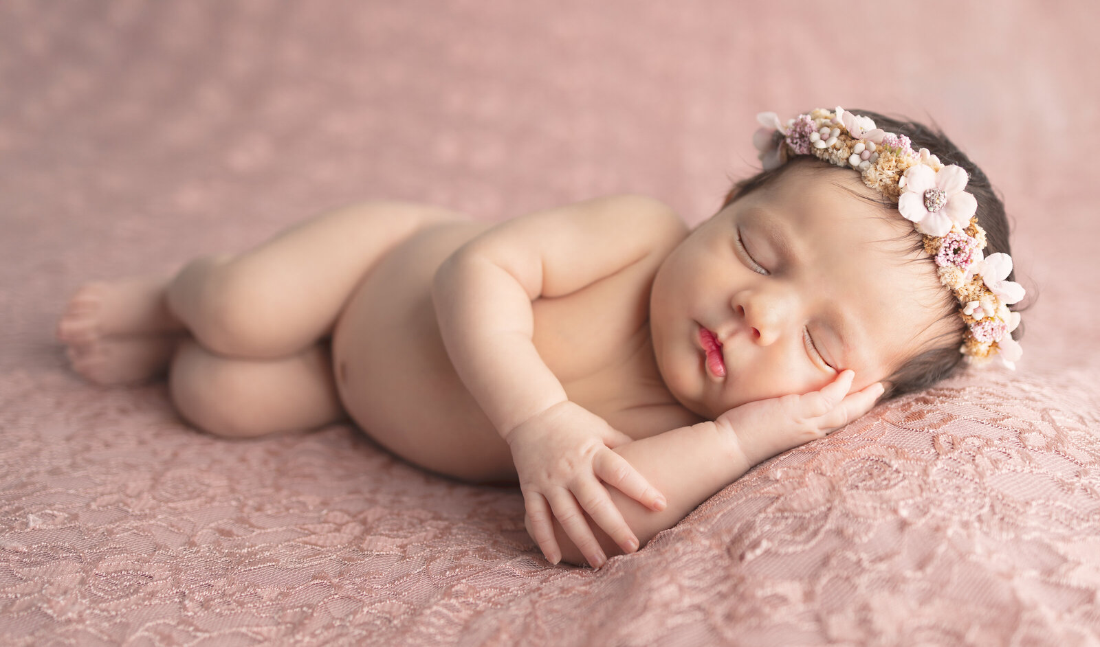 Newborn baby girl with floral headband sleeping on pink lace blanket during professional Waldwick NJ photoshoot