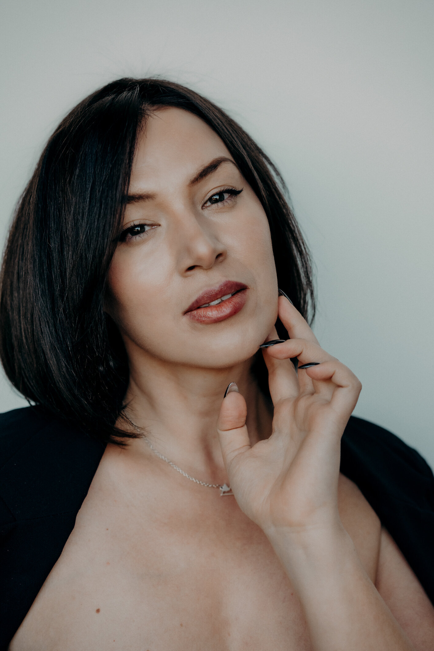 A close-up fine art image of a woman with almost black hair and striking features.  She stares directly at the camera, while tracing her jawline with her well manicured hands.