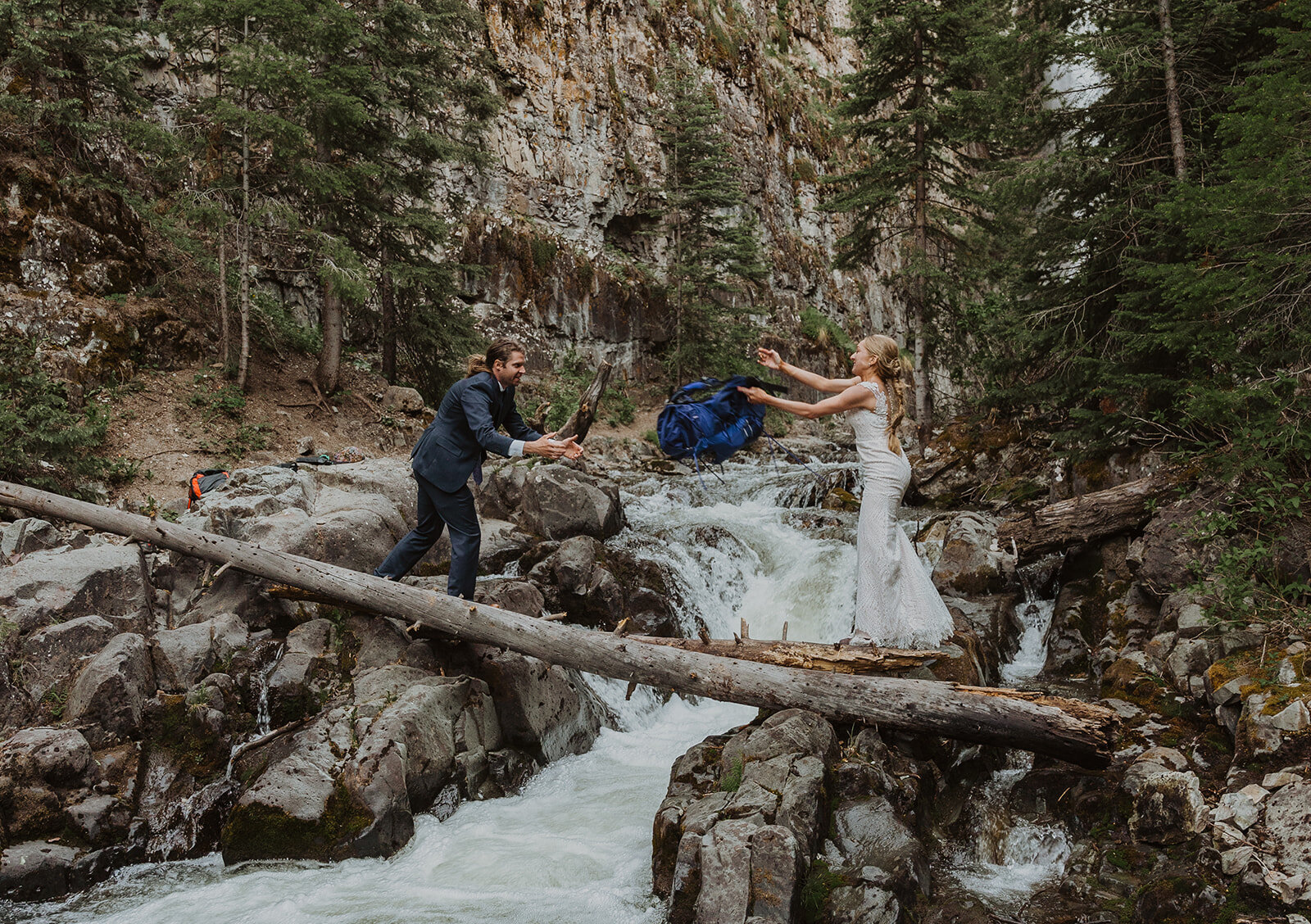 A bride and groom cross a river by delicately walking over a fallen tree . The bride in her dress throws her backpack to the fully dressed groom as the water rushes underneath their feet.
