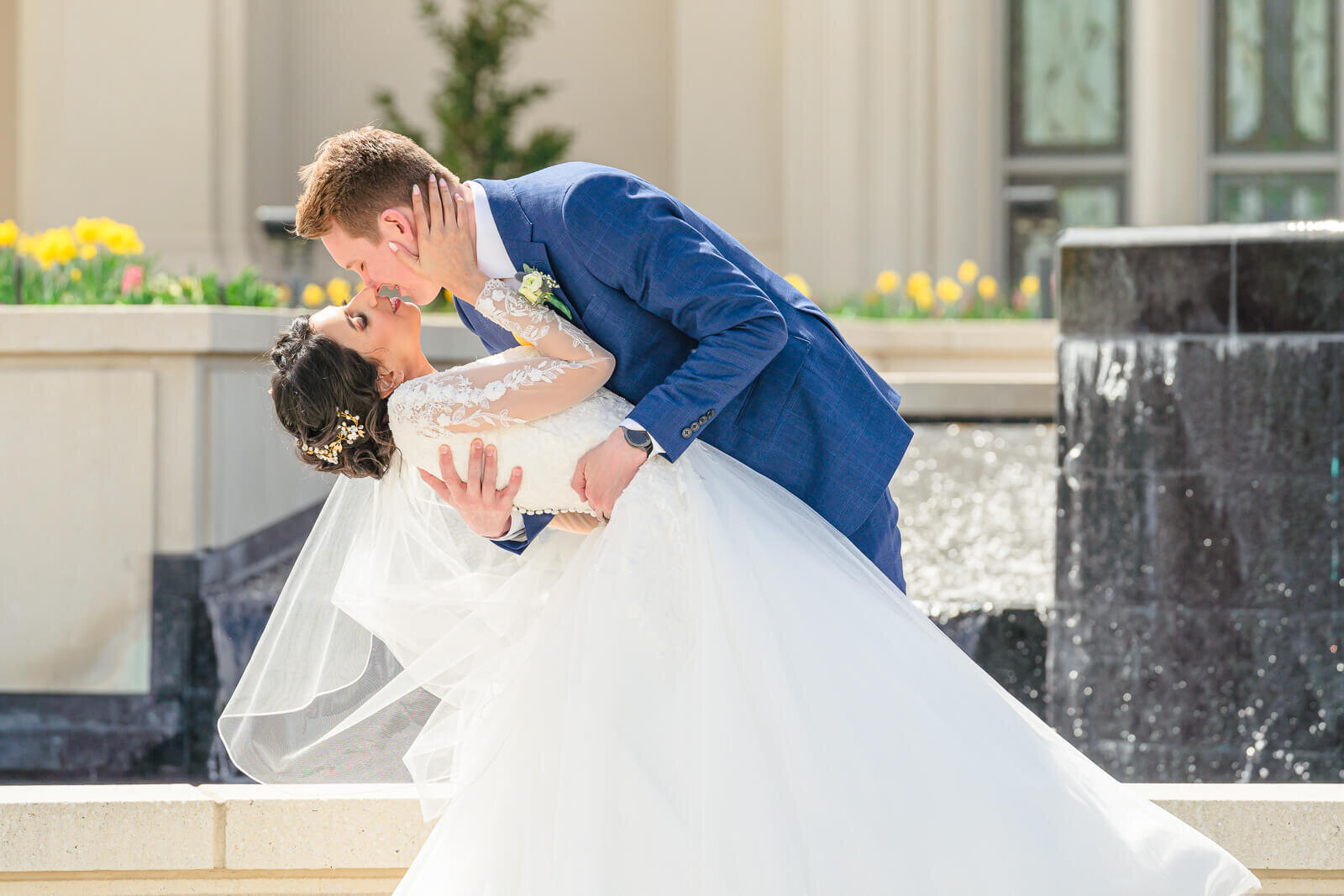 Salt Lake Wedding photography of a groom in a dark blue suit leaning in to kiss his bride in front of the fountain at the Payson Temple
