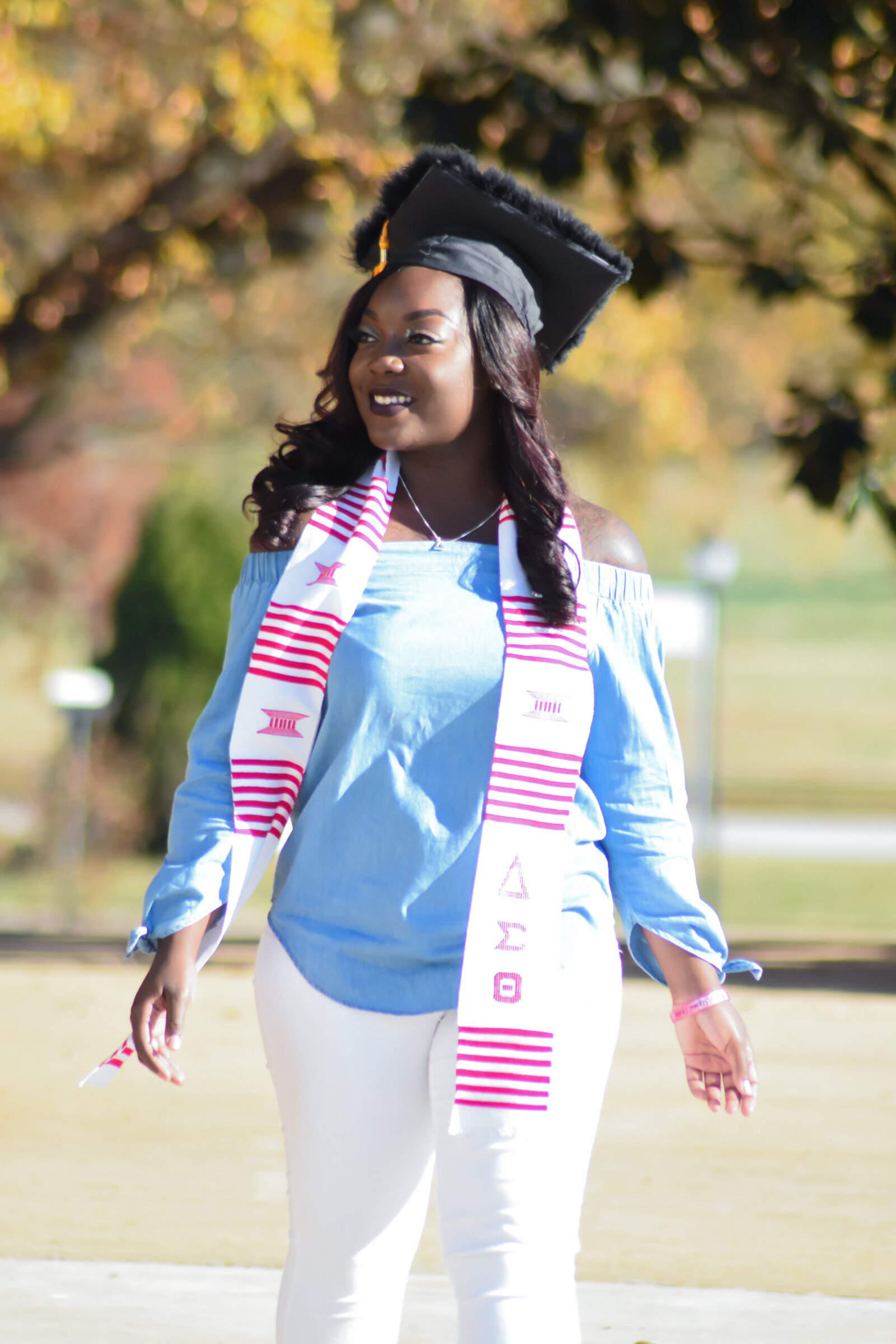 a graduate dressed in a blue shirt and white pants with her graduation stole draped over her shoulders. shes walking down a pathway and wearing a black graduation cap. photographed by Millz Photography in Greenville, SC