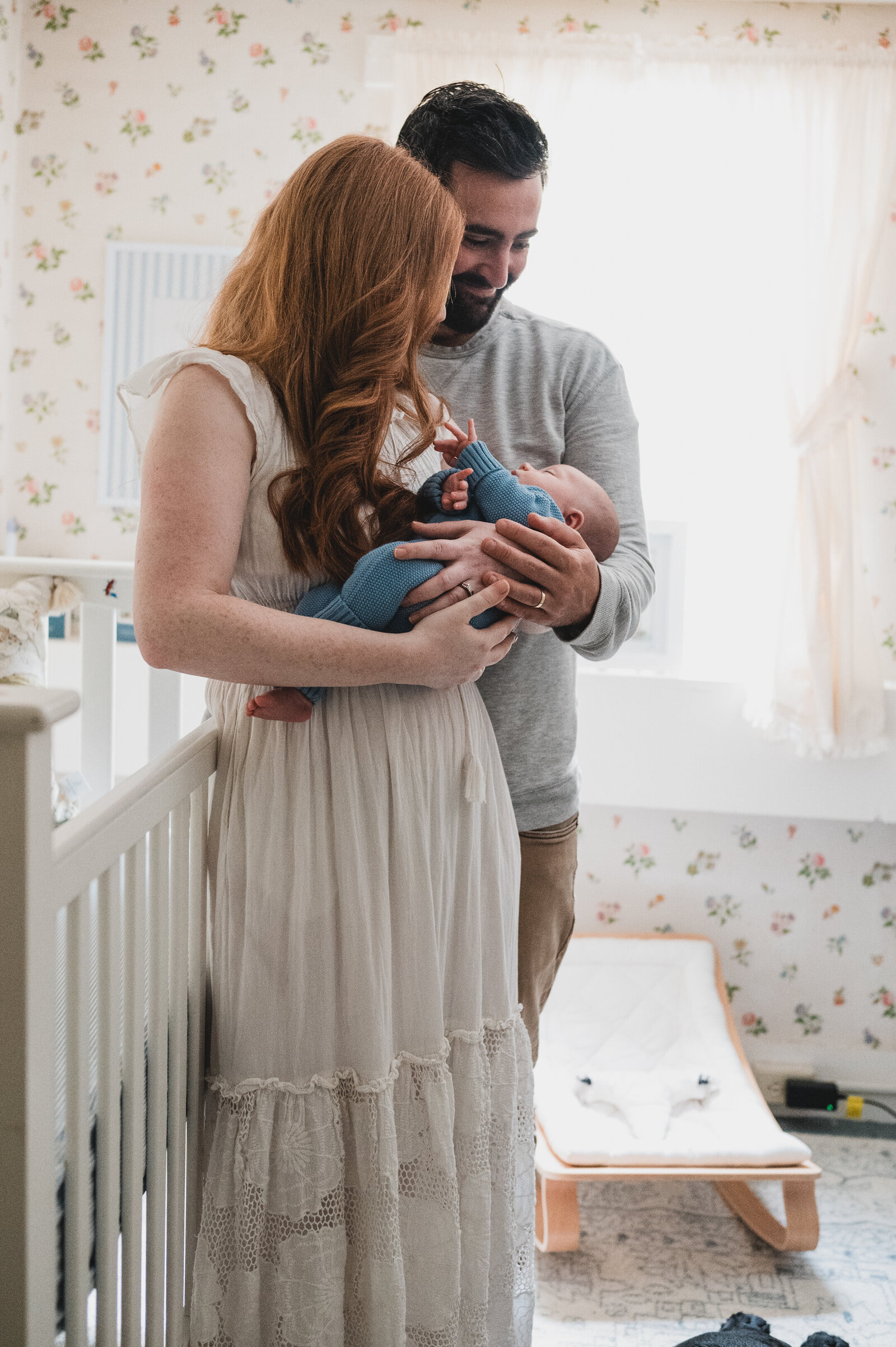 Mother with long red hair holding her newborn baby in a softly lit room, showing a quiet everyday moment