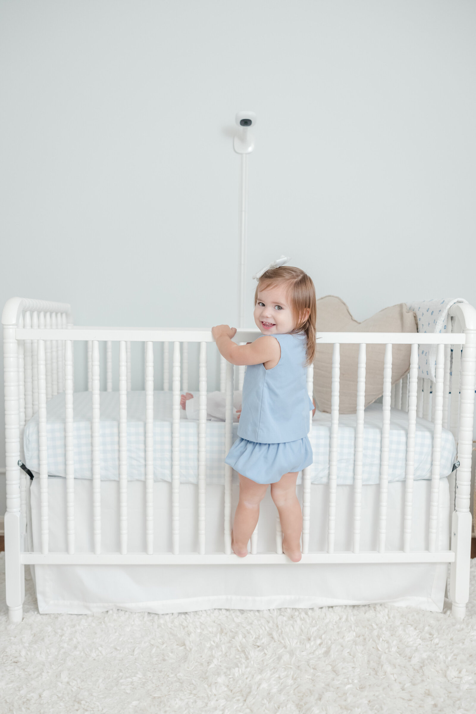 Toddler girl standing on side of a crib and smiling over her shoulder.