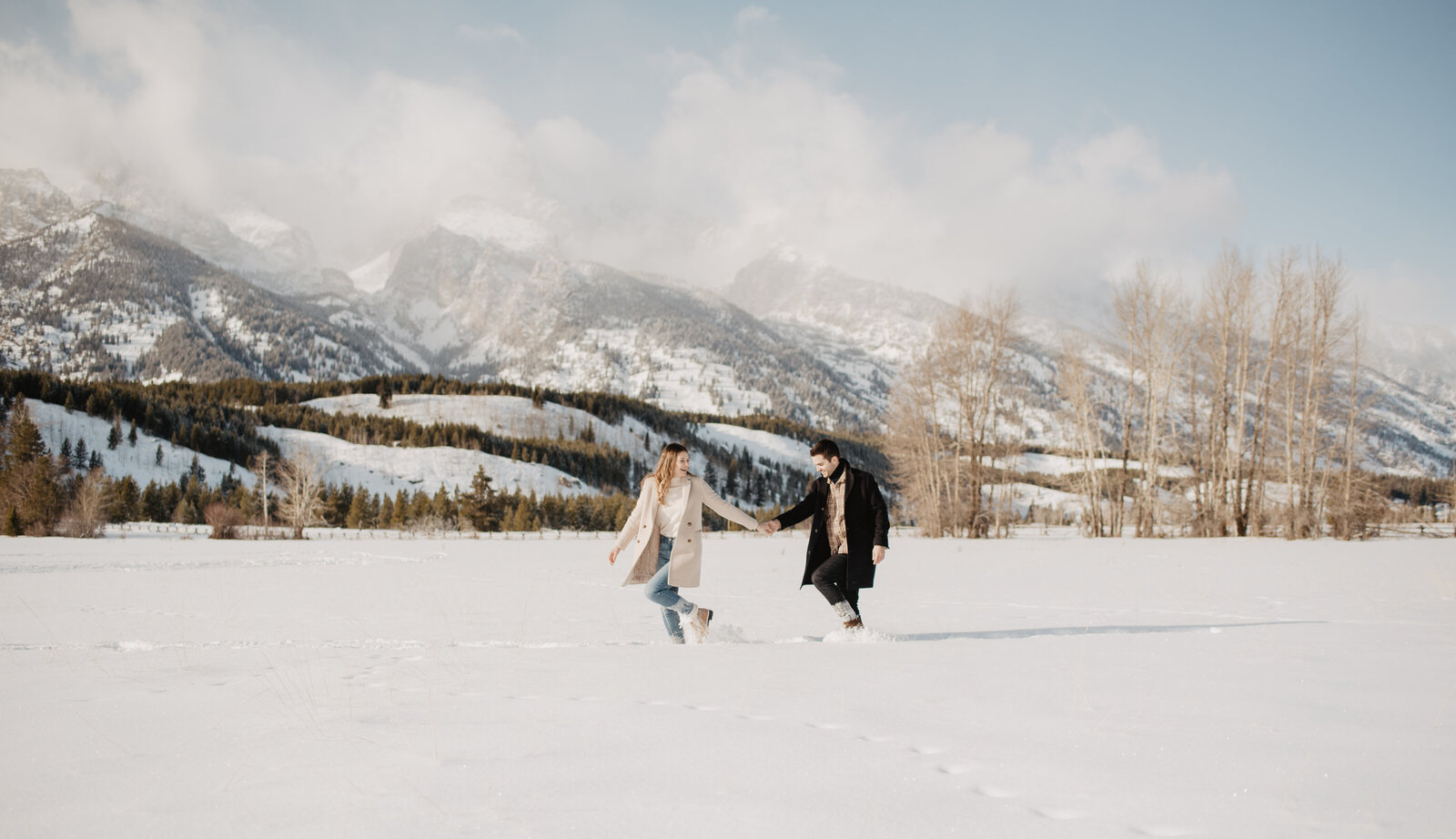 Taggart Lake in the winter with man and woman holding hands and walking through a snowy field with clouds hovering over the snowy mountains photographed by photographers Jackson Hole